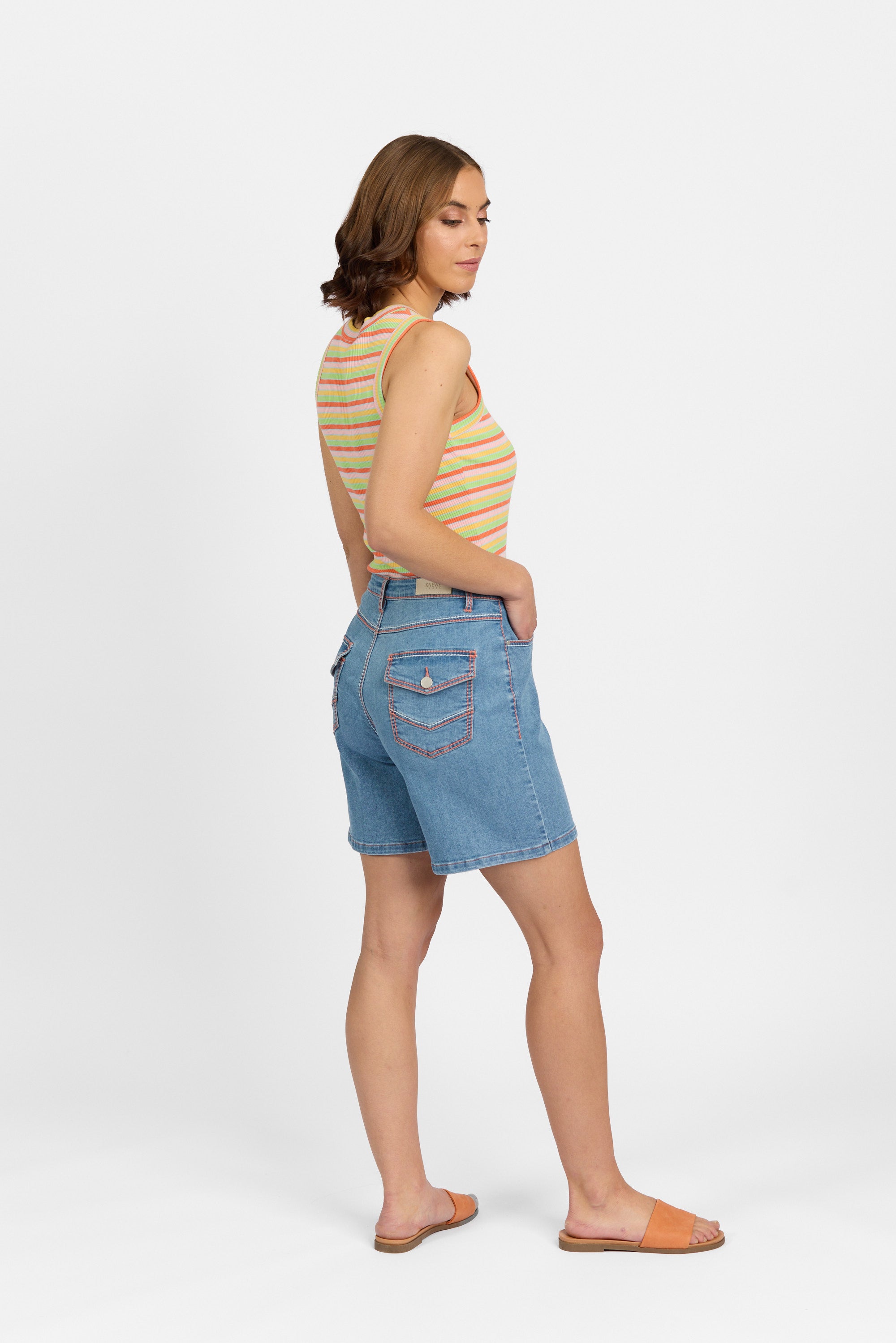 Three-quarter back view of a woman wearing palm blue denim shorts with flap back pockets and contrast stitching, styled with a multicoloured striped tank top and tan slide sandals against a plain light background.