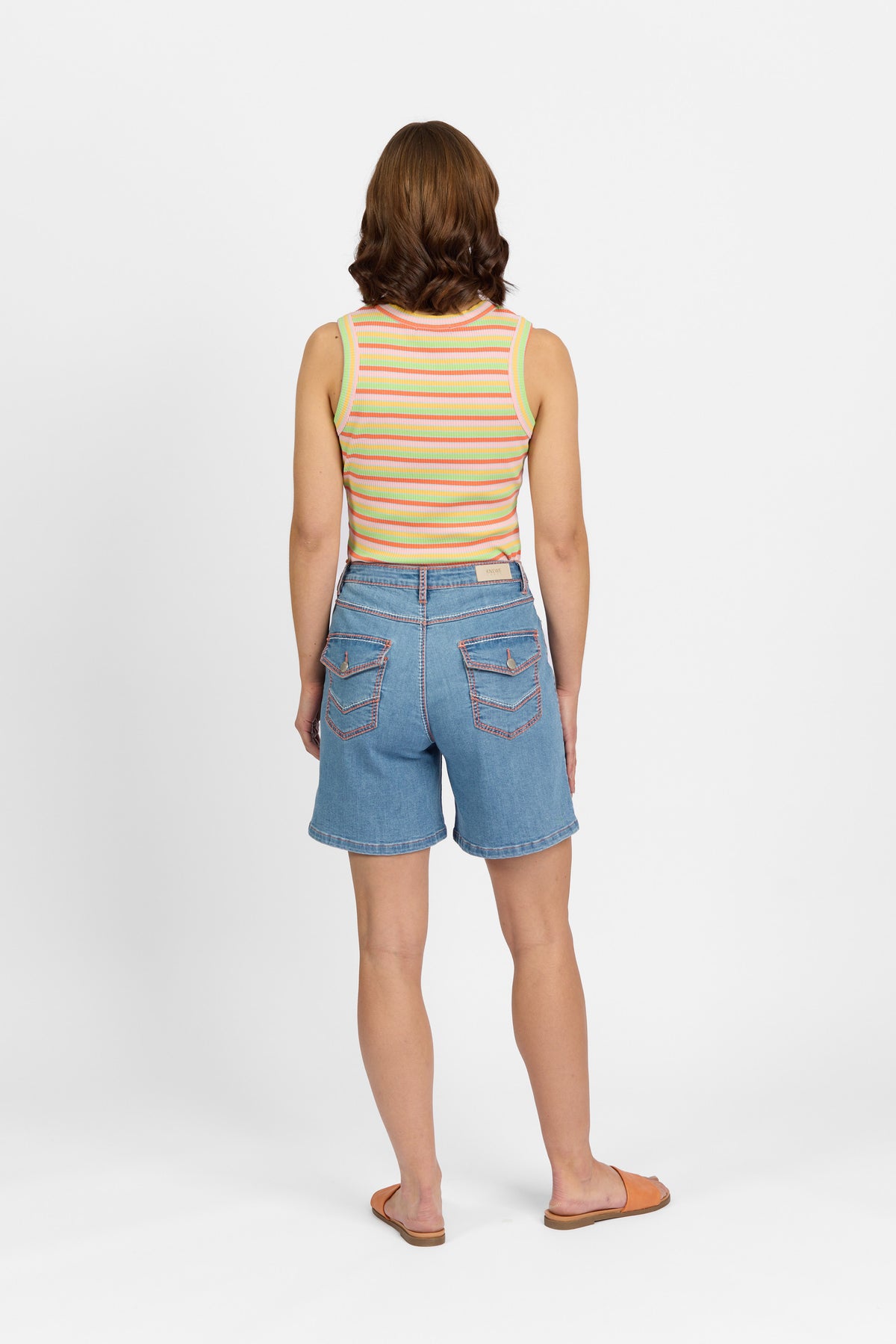 Back view of a woman wearing palm blue denim shorts featuring two flap back pockets with metal button closures and contrast stitching, paired with a multicoloured striped tank top and tan slide sandals.
