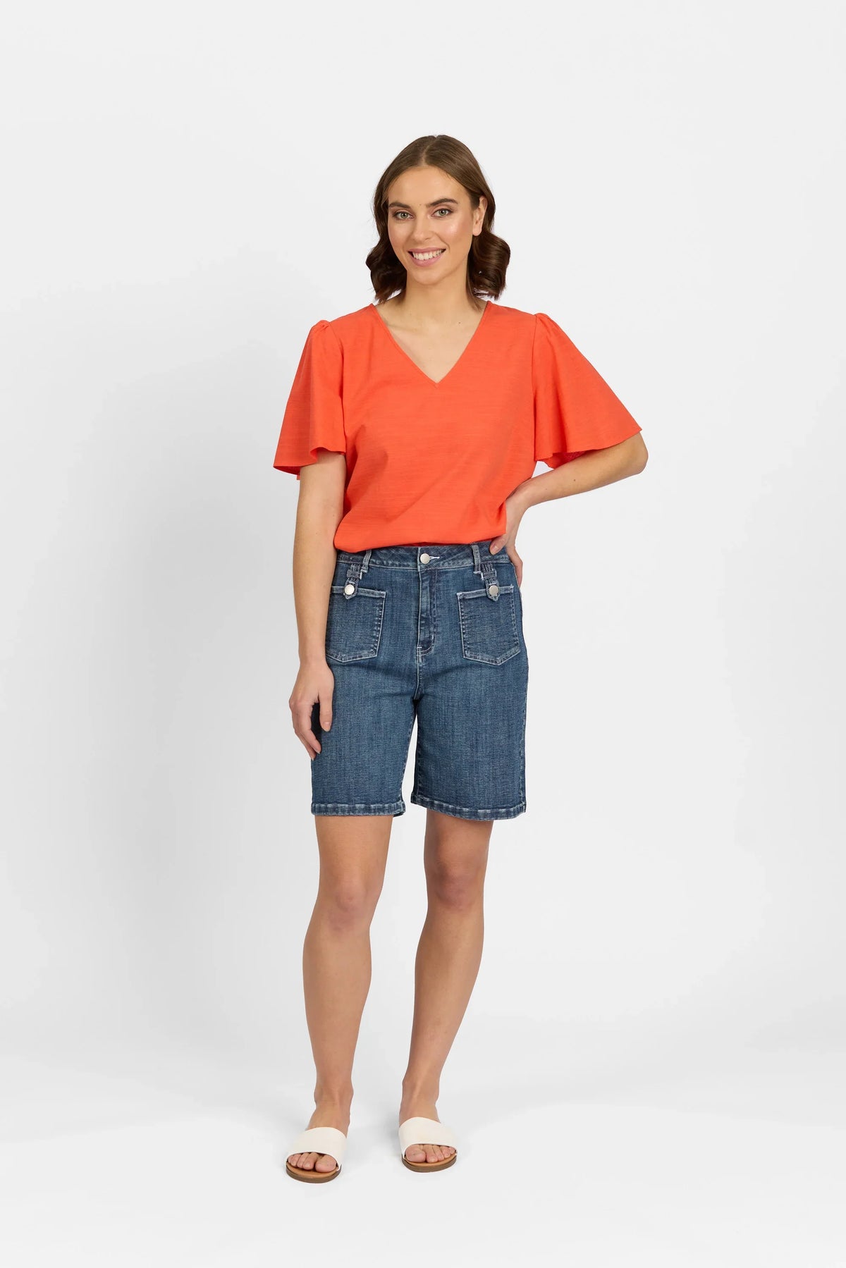 Woman wearing mid-wash denim shorts with front pockets, orange V-neck top, and sandals