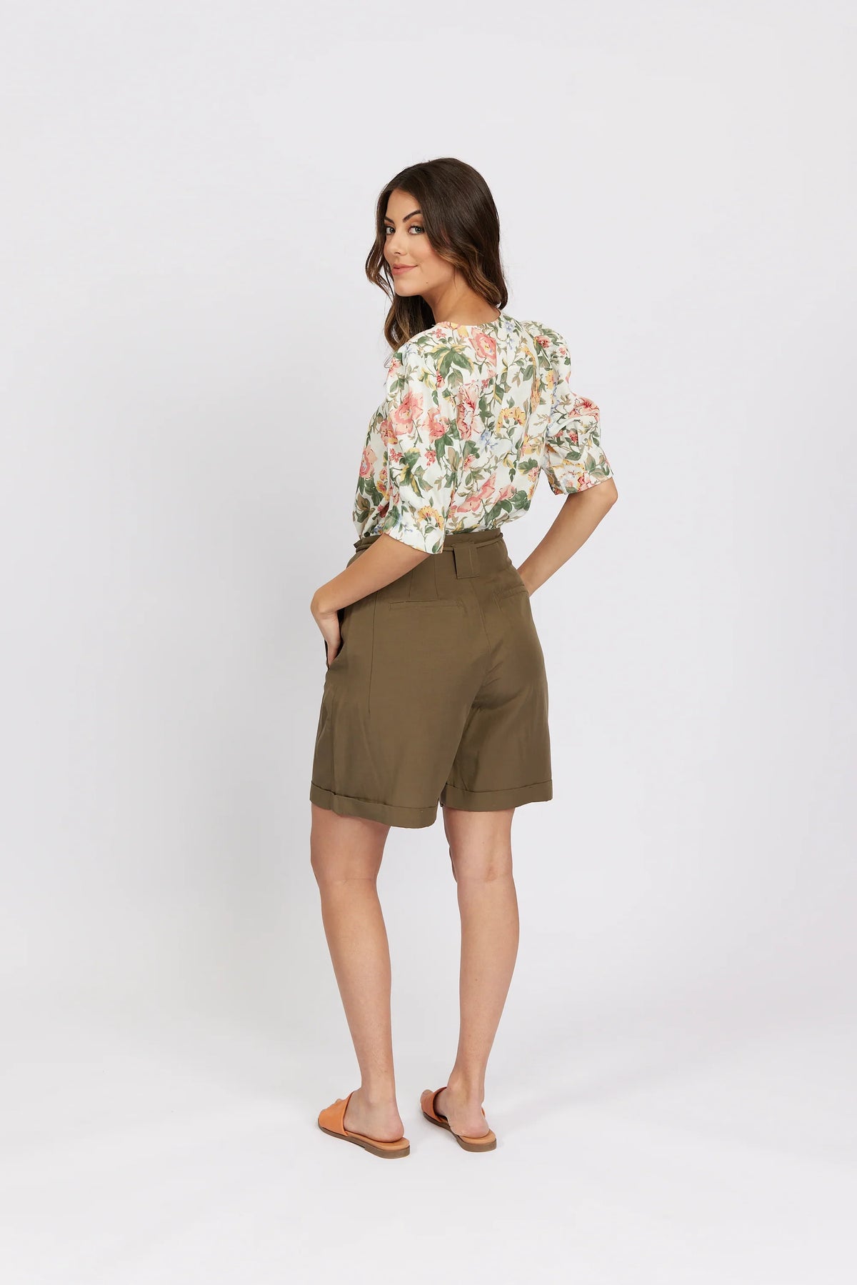 Woman wearing khaki tailored shorts, floral blouse, and sandals, standing on white background