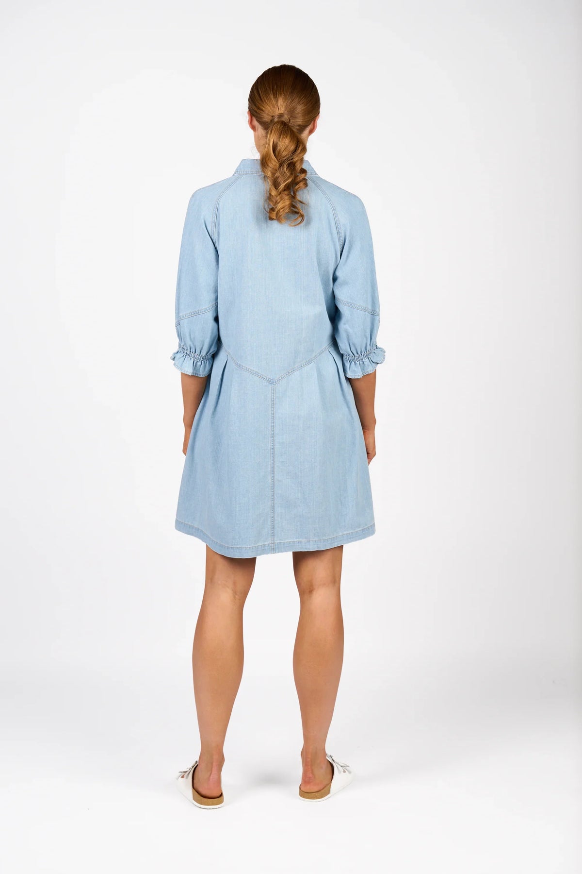 Back view of woman wearing light blue denim dress with puff sleeves, white flat shoes