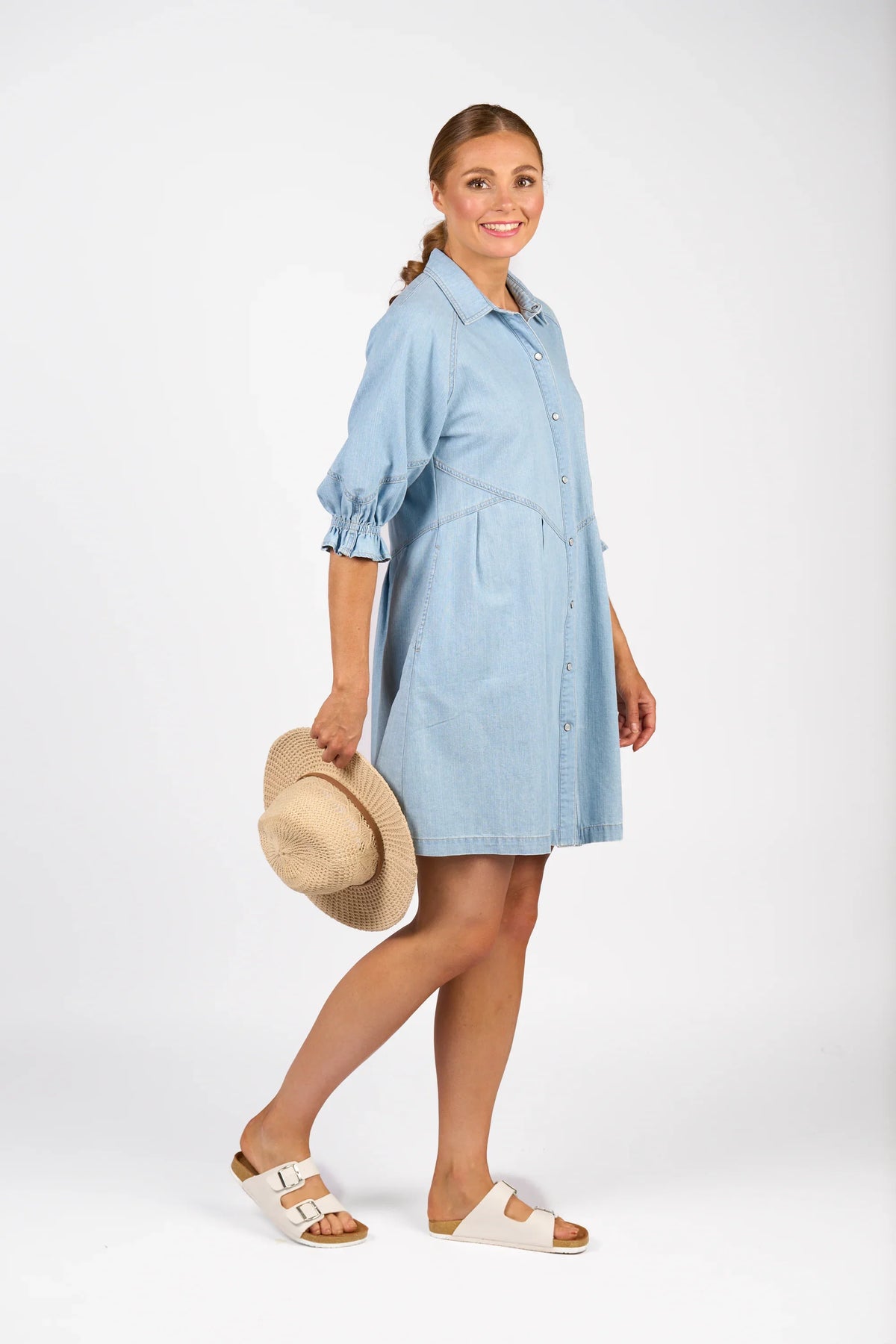 Woman in light wash denim shirt dress with ruffle sleeves, holding straw hat, white sandals