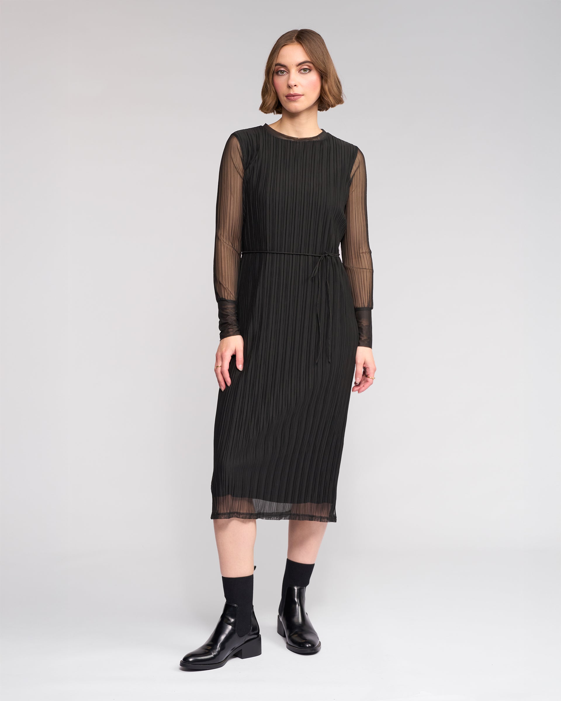 A woman poses against a plain background in the Knewe Label Freddie Dress - Black, featuring sheer long sleeves and vertical pleats, styled with black ankle boots—the ideal slim-fit winter dress for any occasion.