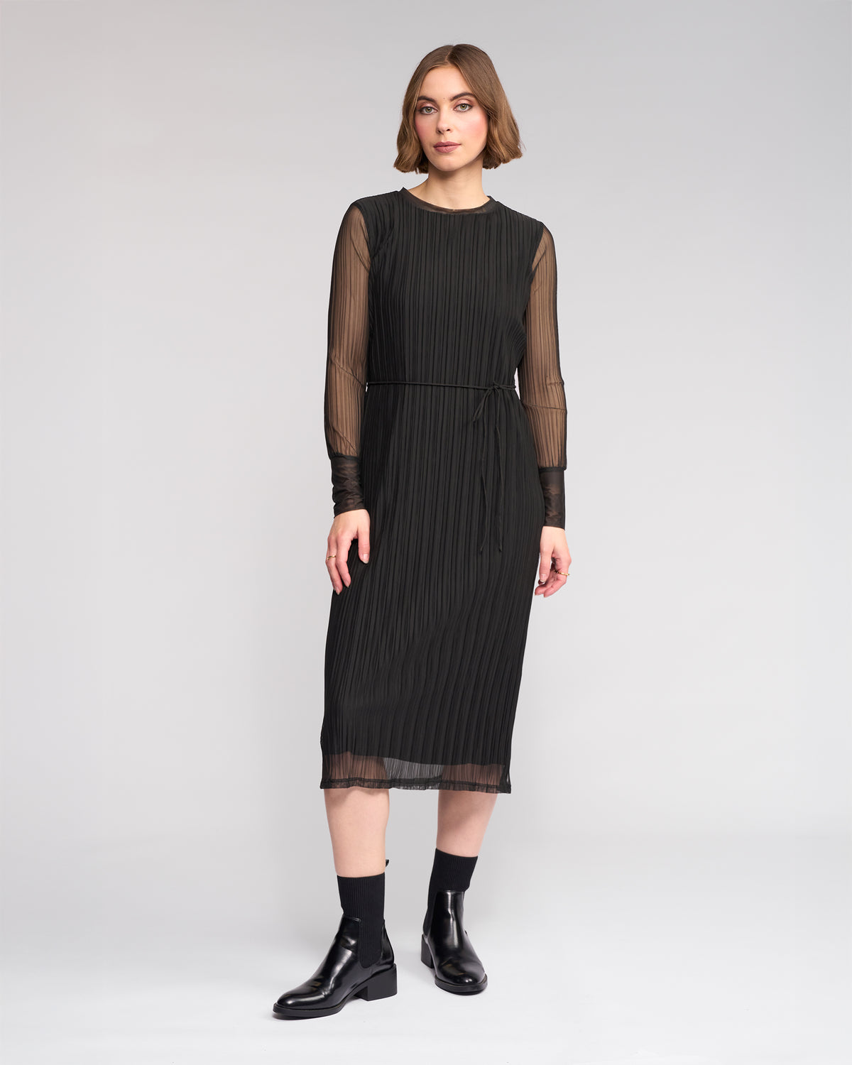 A woman poses against a plain background in the Knewe Label Freddie Dress - Black, featuring sheer long sleeves and vertical pleats, styled with black ankle boots—the ideal slim-fit winter dress for any occasion.