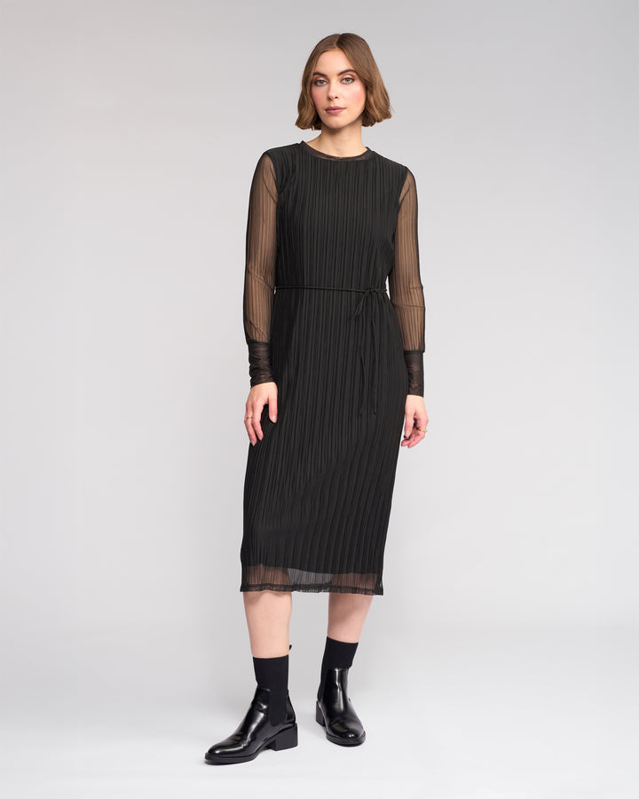 A woman poses against a plain background in the Knewe Label Freddie Dress - Black, featuring sheer long sleeves and vertical pleats, styled with black ankle boots—the ideal slim-fit winter dress for any occasion.