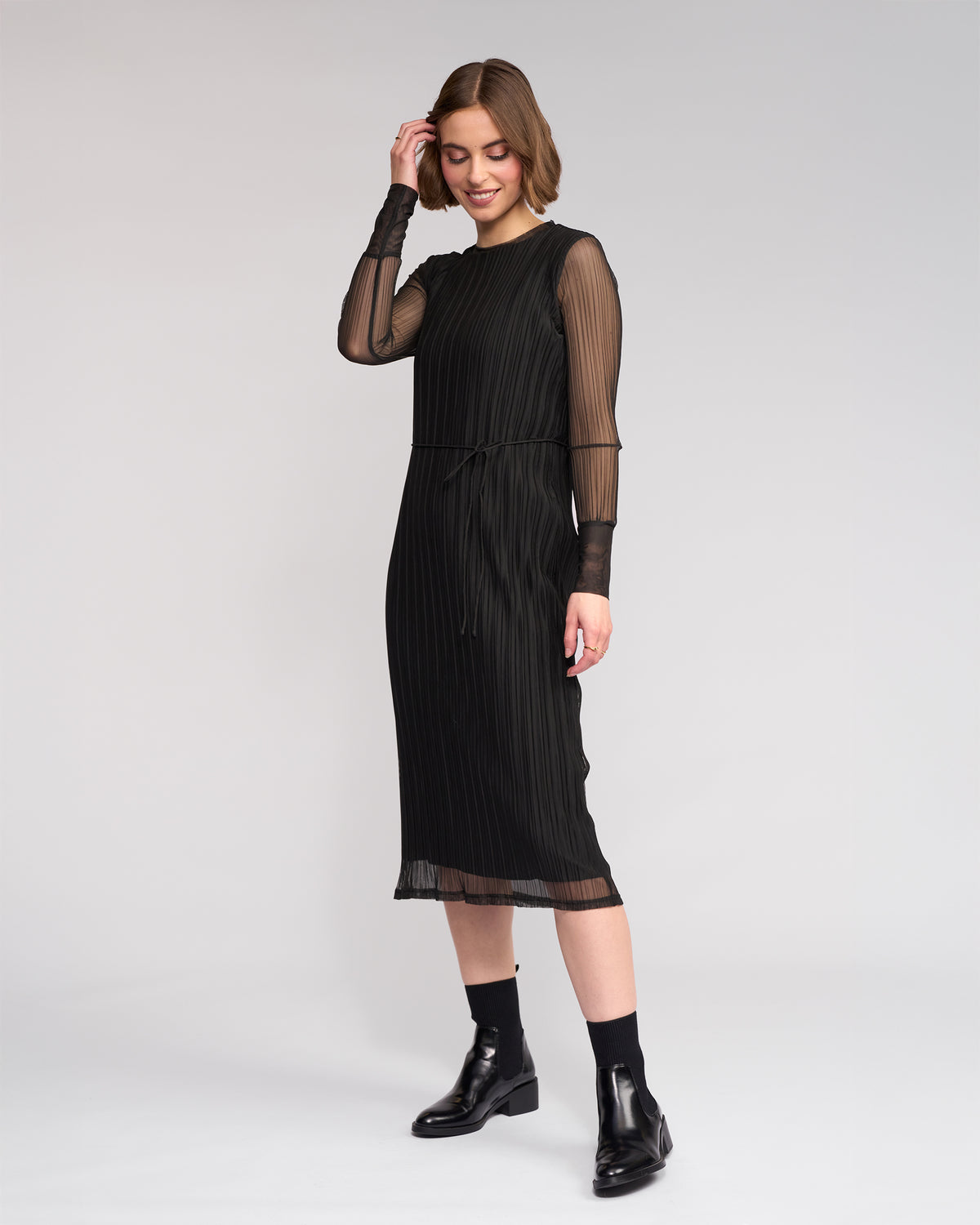 A woman smiles against a plain background, touching her hair as she wears the Knewe Label Freddie Dress - Black—a mesh dress featuring sheer long sleeves and a pleated midi skirt, paired with matching ankle boots for chic winter style.