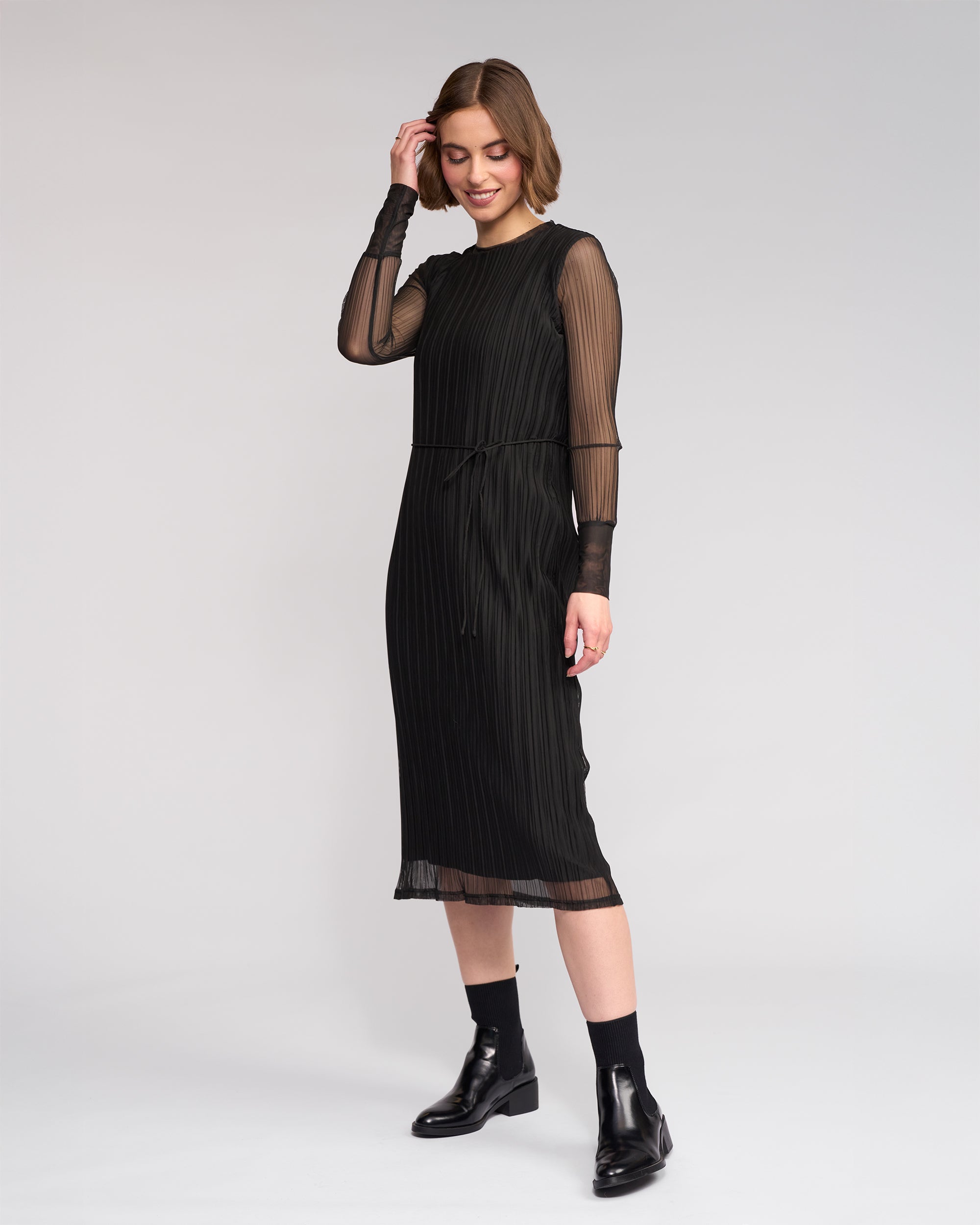 A woman smiles against a plain background, touching her hair as she wears the Knewe Label Freddie Dress - Black—a mesh dress featuring sheer long sleeves and a pleated midi skirt, paired with matching ankle boots for chic winter style.