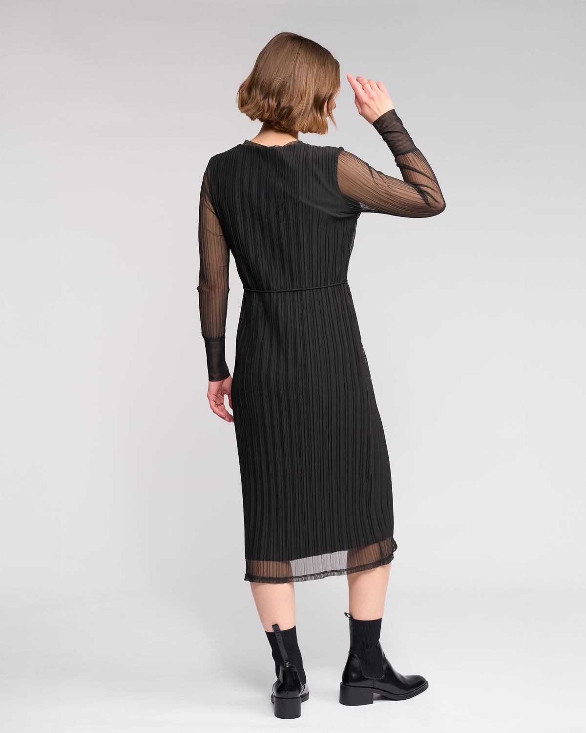 A woman faces away, wearing the Knewe Label Freddie Dress in black—a slim-fit winter dress with sheer sleeves—paired with black ankle boots, set against a plain light background.