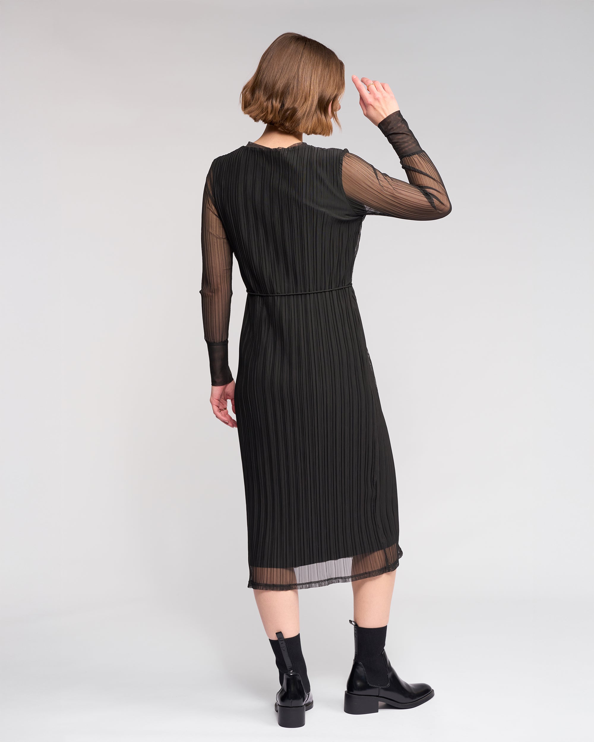 A woman faces away, wearing the Knewe Label Freddie Dress in black—a slim-fit winter dress with sheer sleeves—paired with black ankle boots, set against a plain light background.