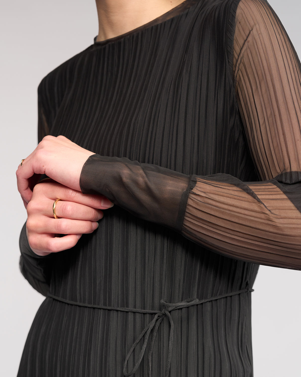 A person wears the Freddie Dress - Black by Knewe Label, a sheer, pleated mesh winter dress with long sleeves, slim fit, thin waist tie, and subtle vertical stripes, accessorized with simple gold rings.