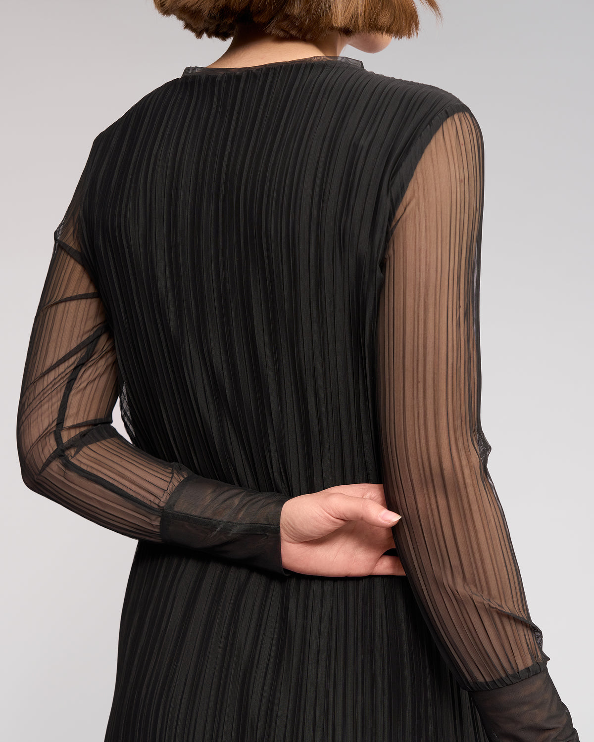 A person with short brown hair is seen from behind wearing the Knewe Label Freddie Dress in black, featuring long sleeves and sheer black mesh, one arm bent to hold the opposite elbow.