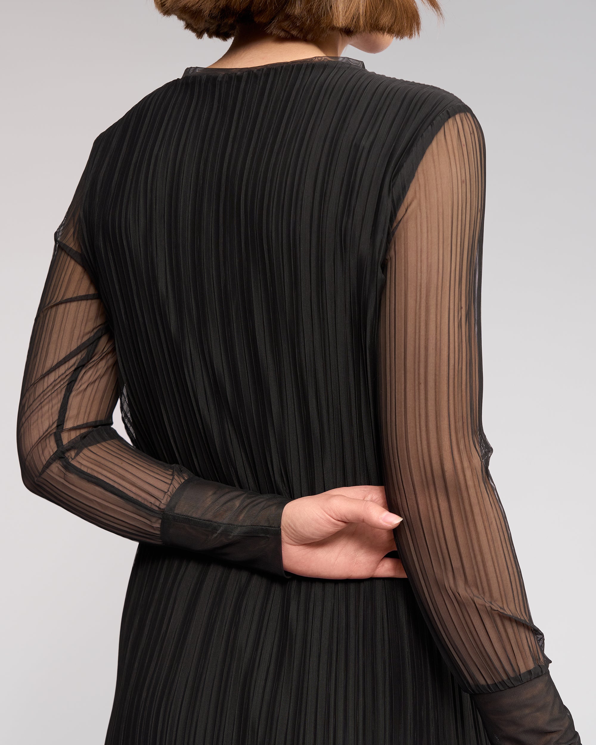 A person with short brown hair is seen from behind wearing the Knewe Label Freddie Dress in black, featuring long sleeves and sheer black mesh, one arm bent to hold the opposite elbow.
