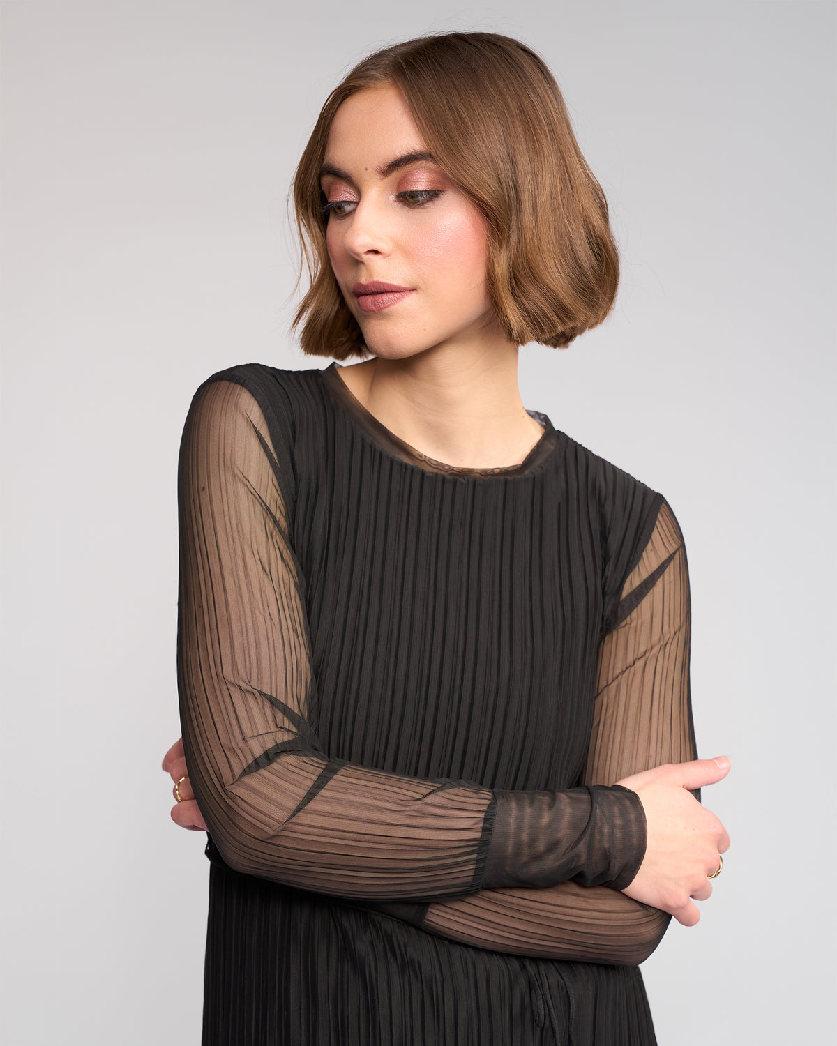 A woman with short light brown hair, wearing the Knewe Label Freddie Dress in black with slim fit and mesh sheer sleeves, stands with arms crossed and looks to her left against a plain light background.