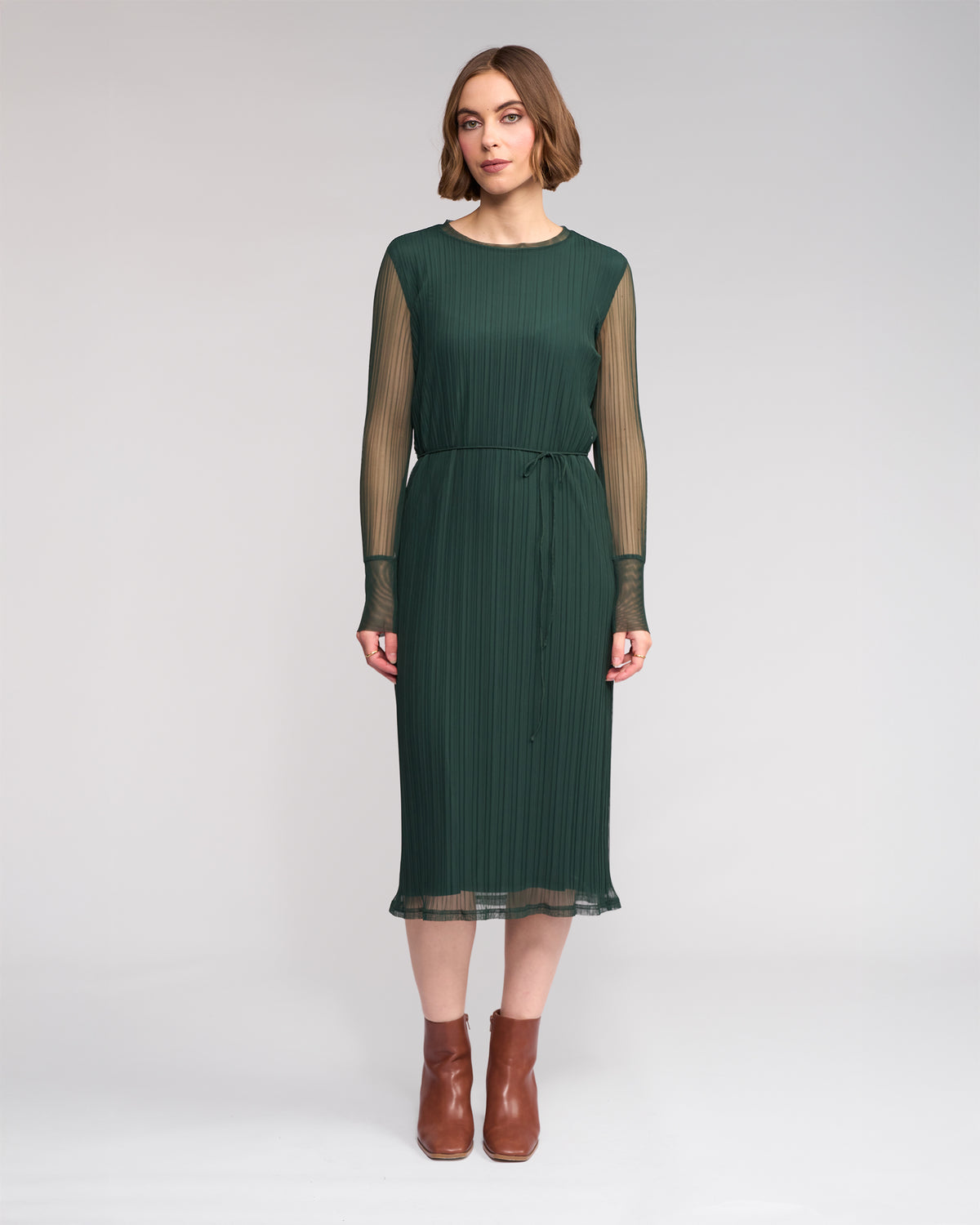 A woman faces forward in the Freddie Dress - Hunter by Knewe Label, featuring chic green mesh with sheer long sleeves and a slim waist tie, paired with brown ankle boots. She poses against a plain light background, ideal for winter style.
