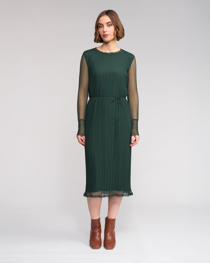 A woman faces forward in the Freddie Dress - Hunter by Knewe Label, featuring chic green mesh with sheer long sleeves and a slim waist tie, paired with brown ankle boots. She poses against a plain light background, ideal for winter style.