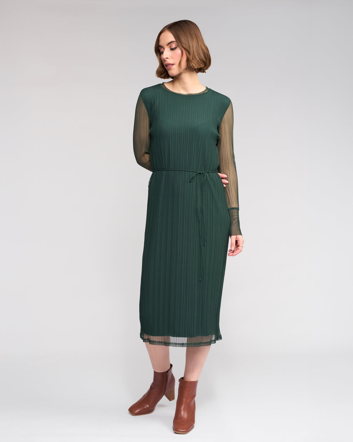 A woman wearing the Knewe Label Freddie Dress - Hunter, a long sleeve green mesh dress with sheer sleeves and a thin waist tie, paired with brown ankle boots, stands against a plain light background.
