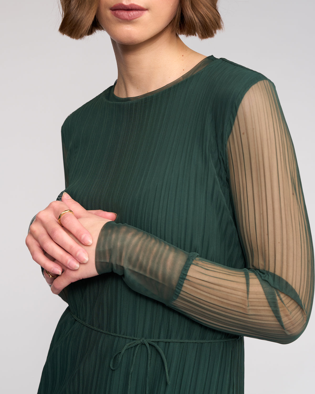A woman with short brown hair models the Knewe Label Freddie Dress - Hunter, featuring sheer, pleated long sleeves. She clasps her hands together, with only the lower part of her face visible against a plain neutral background.
