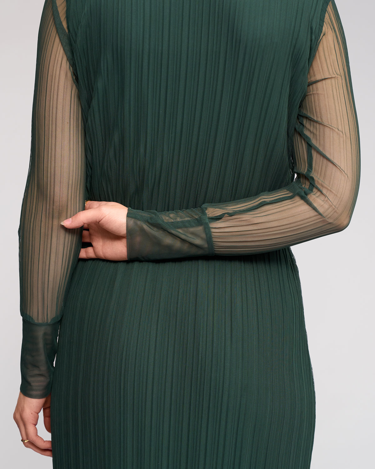 Wearing the Knewe Label Freddie Dress - Hunter, a person stands with their back to the camera in a sheer, long sleeve green mesh dress, left arm bent and holding their right elbow.