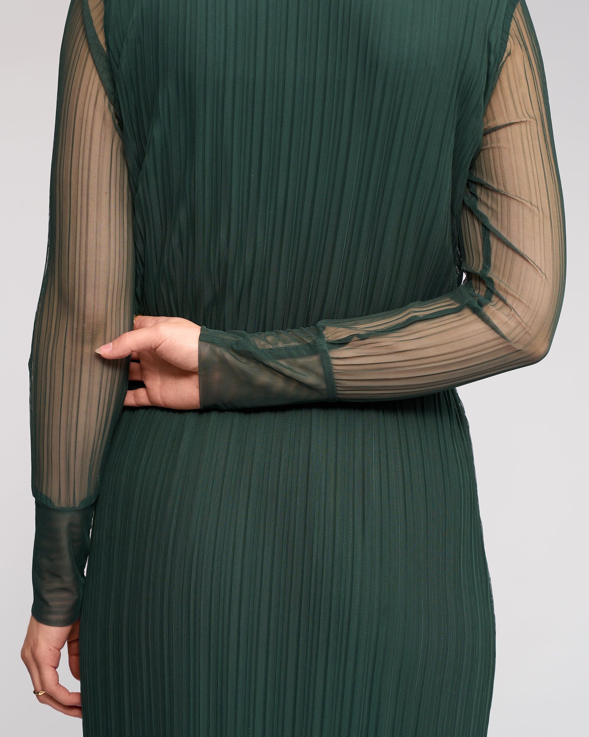 Wearing the Knewe Label Freddie Dress - Hunter, a person stands with their back to the camera in a sheer, long sleeve green mesh dress, left arm bent and holding their right elbow.