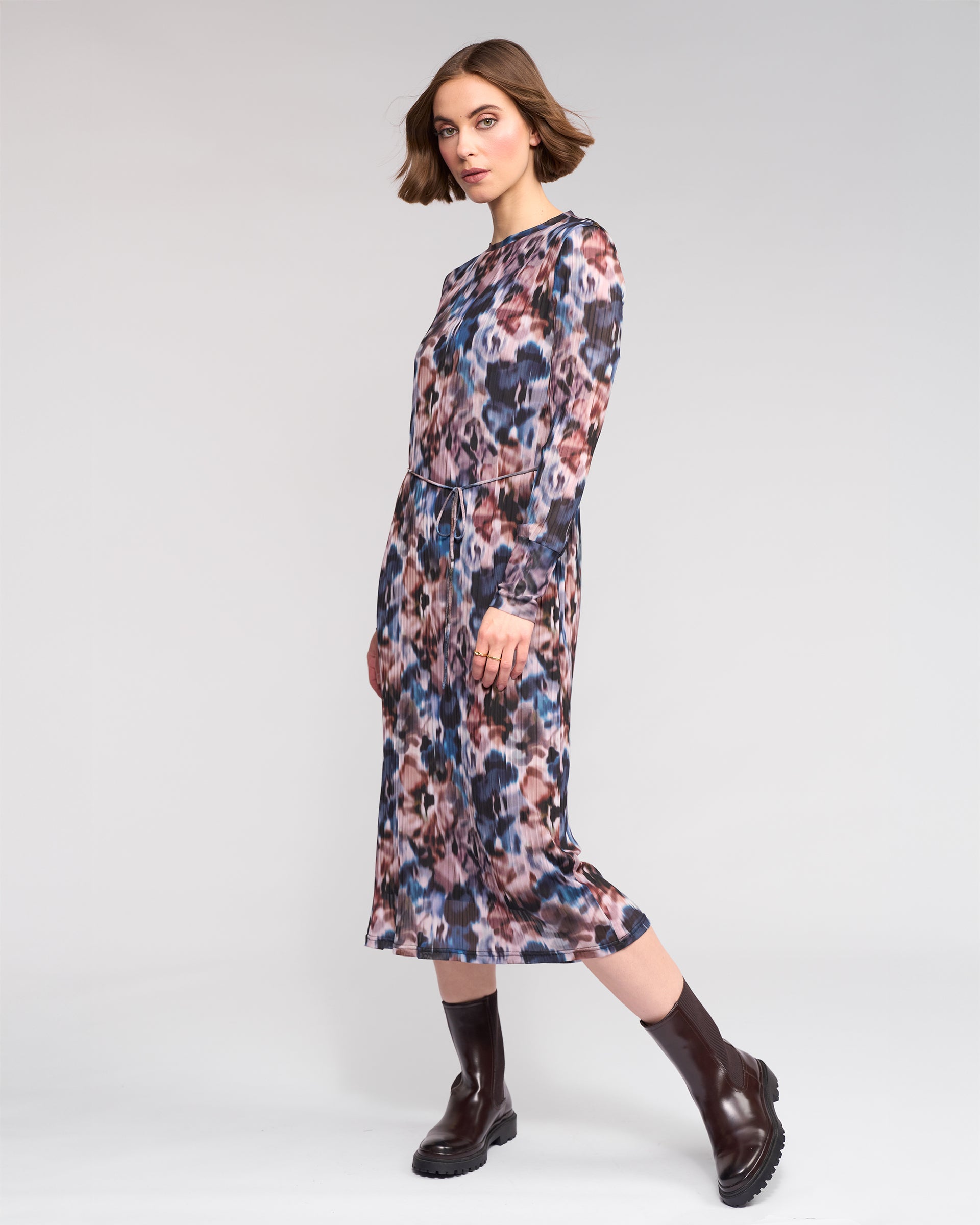 A woman with short brown hair models the Knewe Label Freddie Dress - Celine, a long sleeve abstract print dress in pink, blue, and brown hues, paired with dark brown ankle boots against a plain light background.