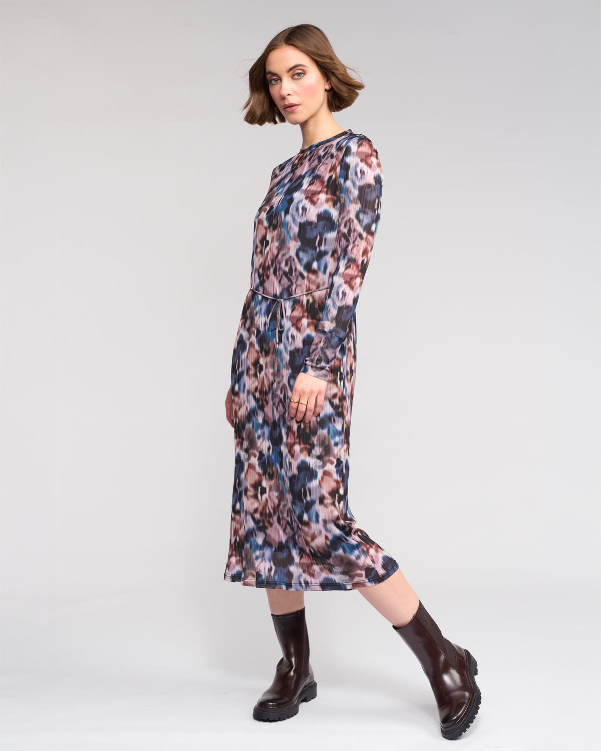 A woman with short brown hair models the Knewe Label Freddie Dress - Celine, a long sleeve abstract print dress in pink, blue, and brown hues, paired with dark brown ankle boots against a plain light background.