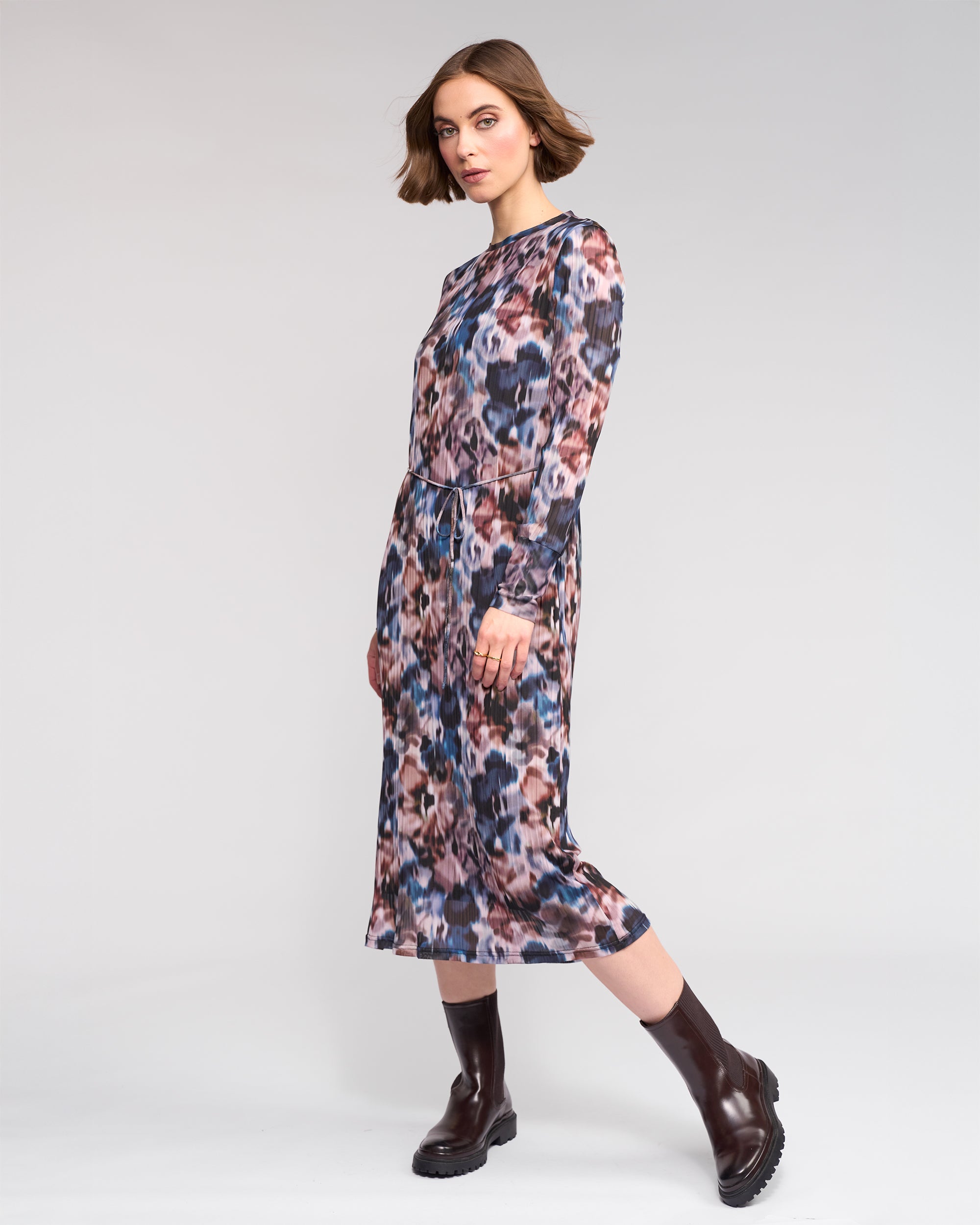 A woman with short brown hair models the Knewe Label Freddie Dress - Celine, a long sleeve abstract print dress in pink, blue, and brown hues, paired with dark brown ankle boots against a plain light background.