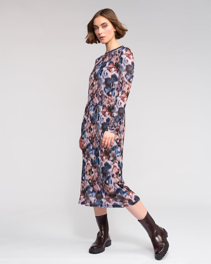 A woman with short brown hair models the Knewe Label Freddie Dress - Celine, a long sleeve abstract print dress in pink, blue, and brown hues, paired with dark brown ankle boots against a plain light background.