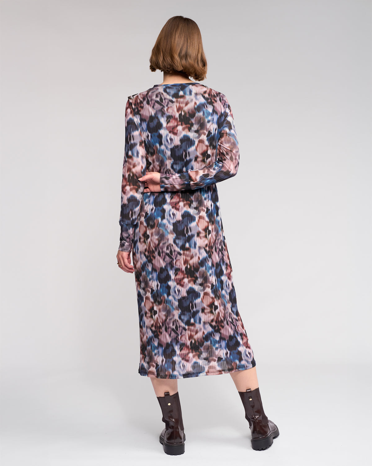 A person with short brown hair faces away, wearing the Knewe Label Freddie Dress - Celine, featuring an abstract print, paired with brown ankle boots against a plain light background.