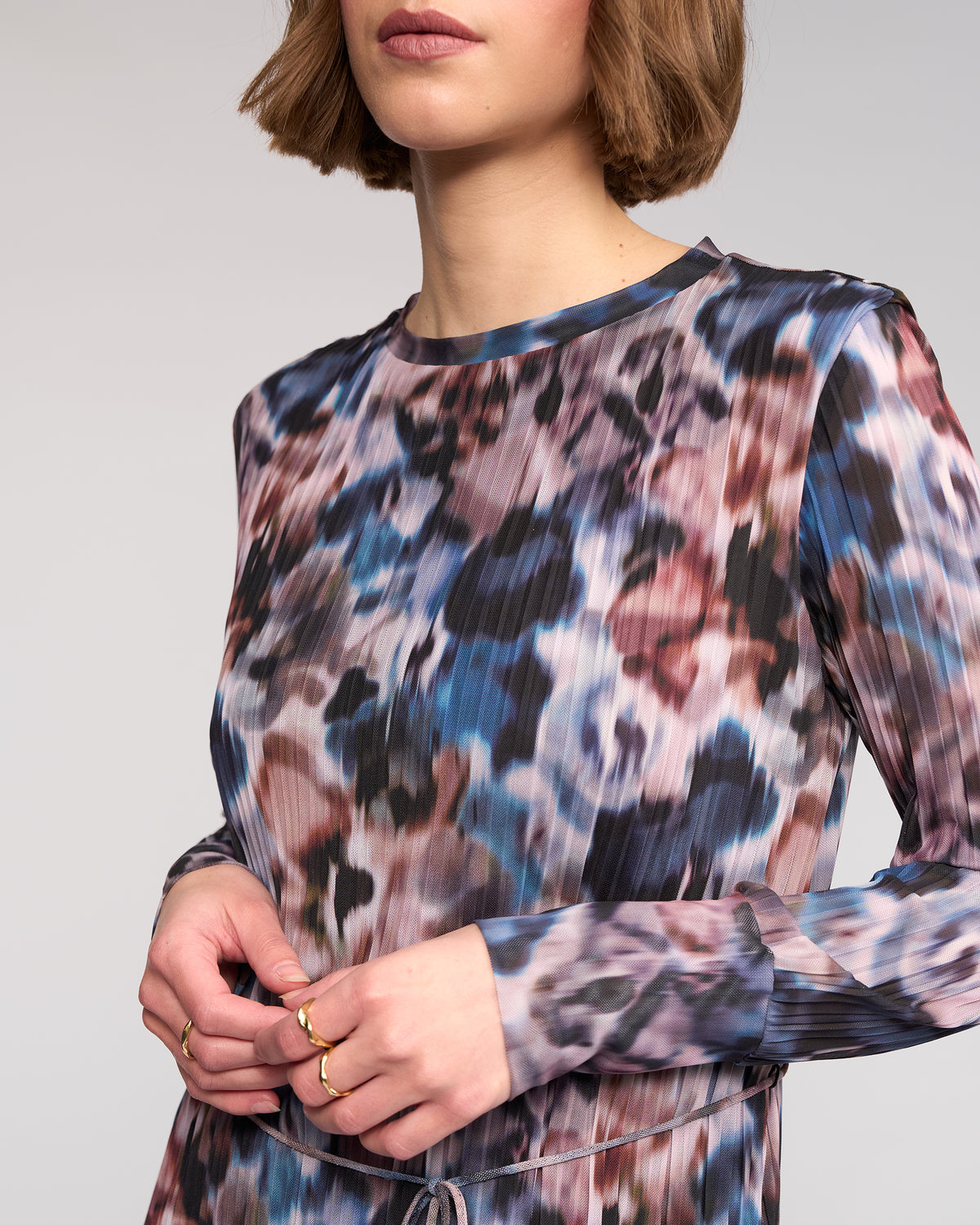 A woman with short brown hair wears the Knewe Label Freddie Dress - Celine, a long sleeve abstract print winter dress in blue, purple, and brown. She accessorizes with gold rings. The image is cropped at her shoulders and midsection.