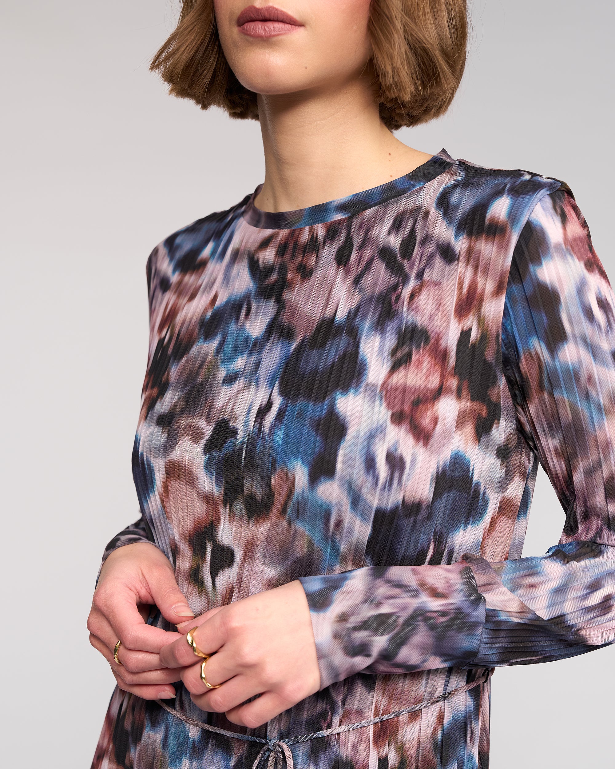 A woman with short brown hair wears the Knewe Label Freddie Dress - Celine, a long sleeve abstract print winter dress in blue, purple, and brown. She accessorizes with gold rings. The image is cropped at her shoulders and midsection.