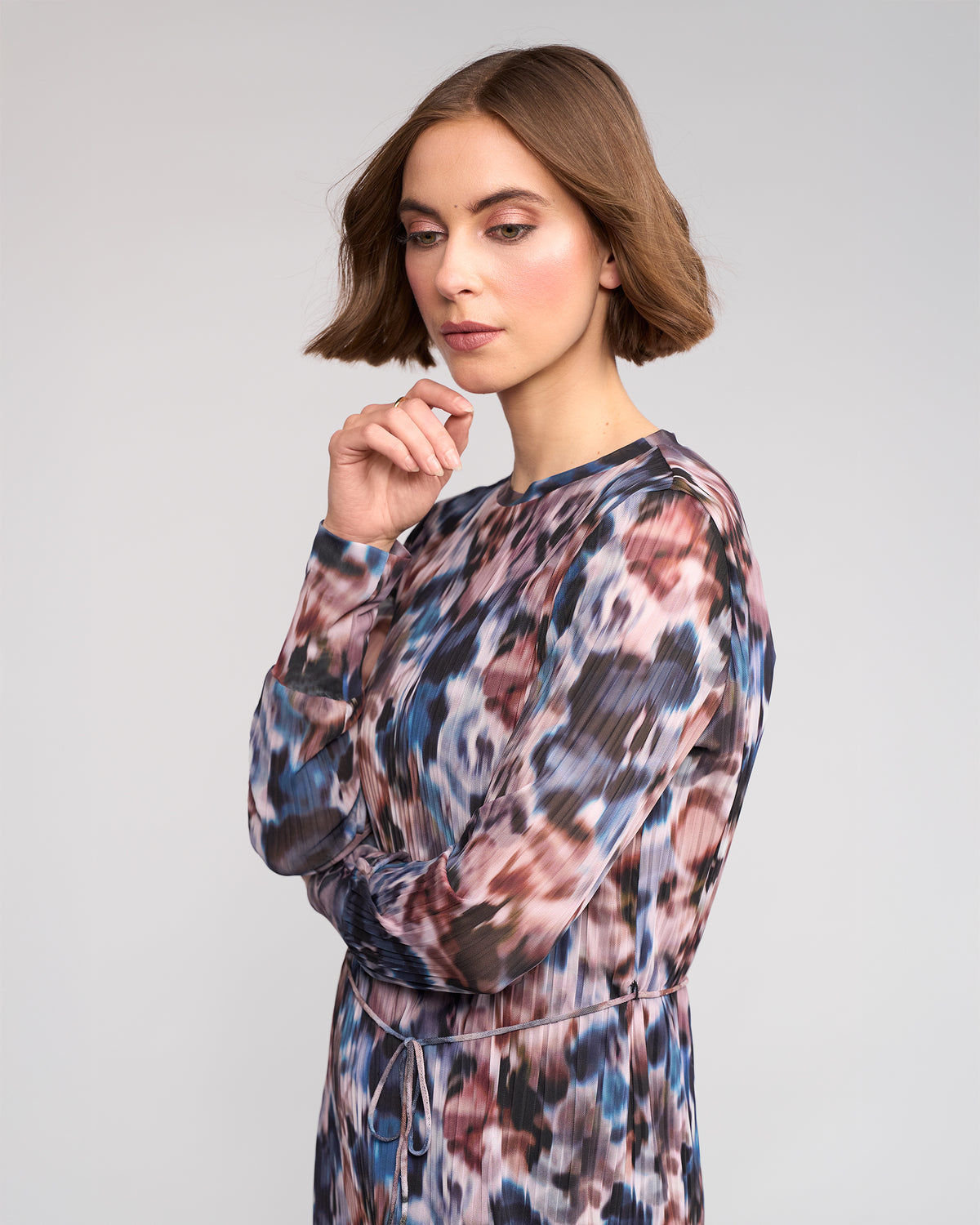 A woman with short brown hair stands in profile, wearing the Knewe Label Freddie Dress - Celine, a long-sleeved winter dress with an abstract print. She gazes down thoughtfully, resting her chin on her hand against a neutral background.