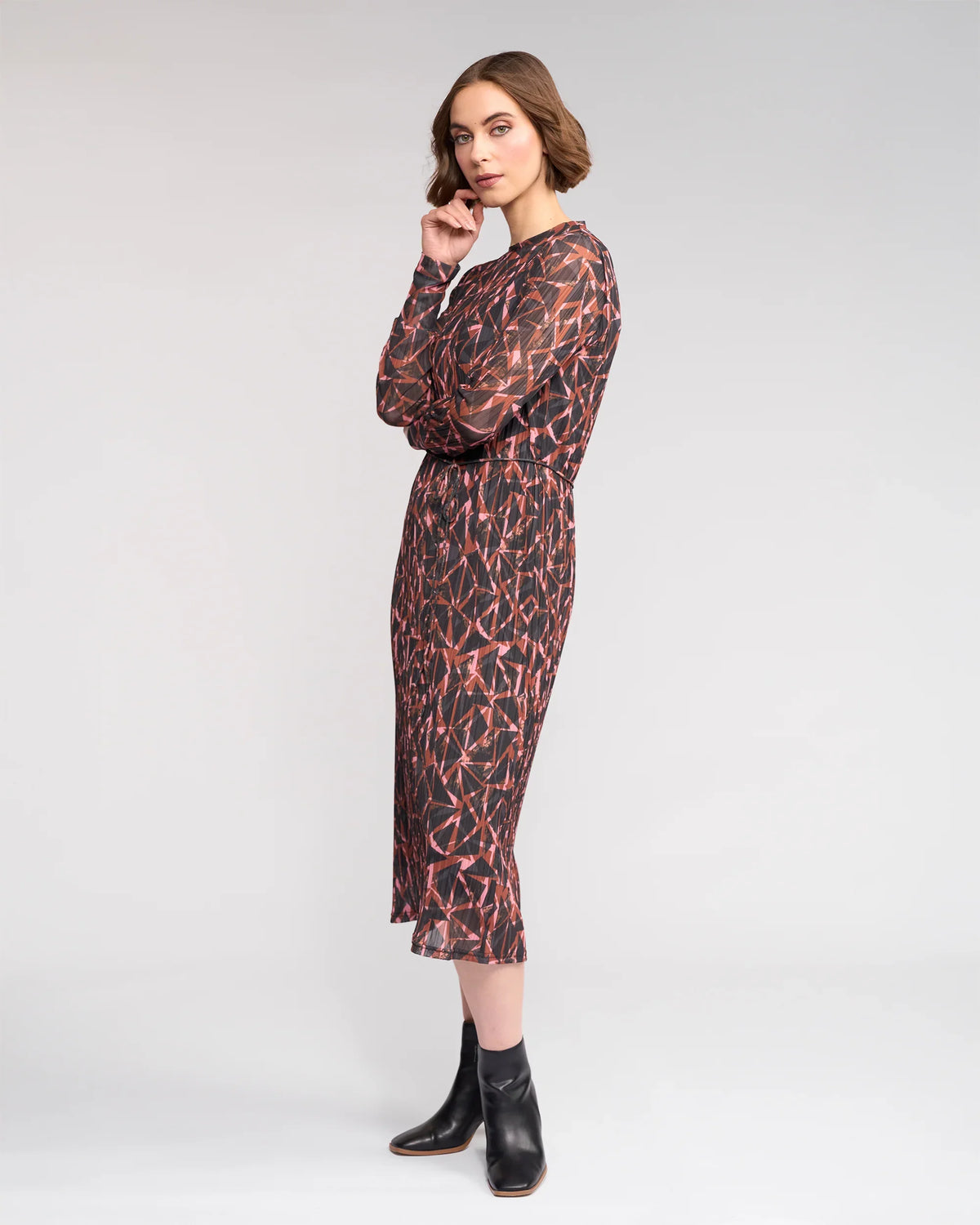 A woman stands sideways wearing the Knewe Label Freddie Dress - Dusk, a long-sleeved, knee-length winter dress with a red and black print, paired with black ankle boots against a plain light gray background.