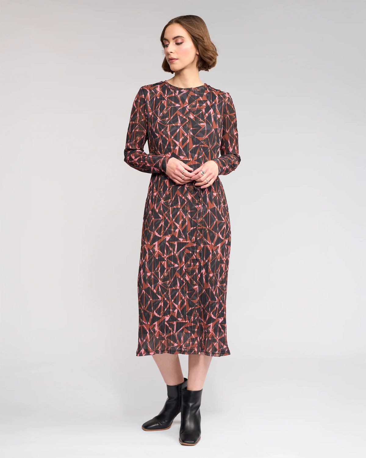 A woman stands against a plain light gray background, wearing the Knewe Label Freddie Dress - Dusk, a long-sleeve, midi winter dress in red and black crinkle mesh, paired with black ankle boots. She looks slightly to the side with hands together in front.