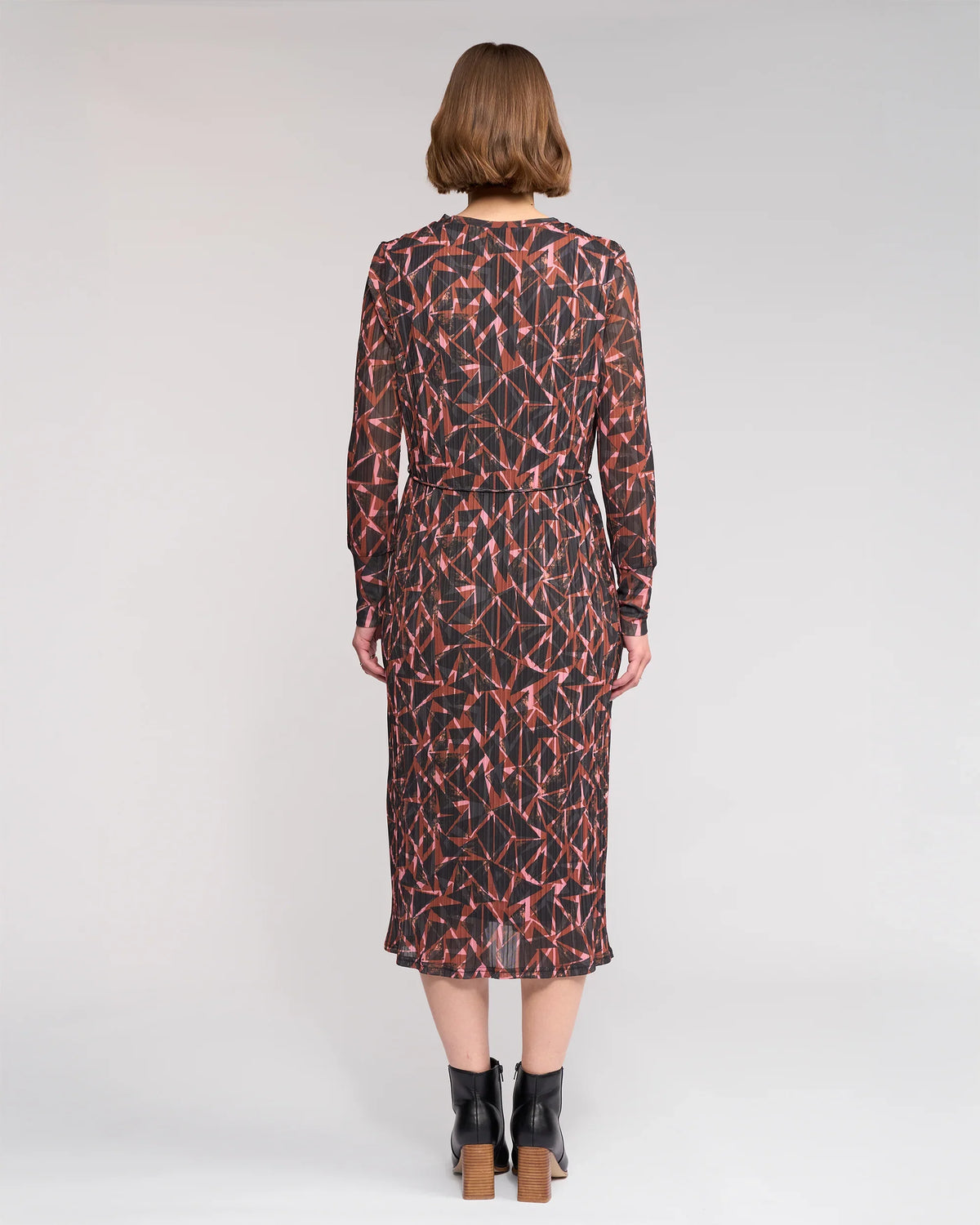 A woman with short light brown hair wears the Knewe Label Freddie Dress - Dusk, a long-sleeved, mid-length winter dress in red and black, paired with black block-heel ankle boots against a plain light background.
