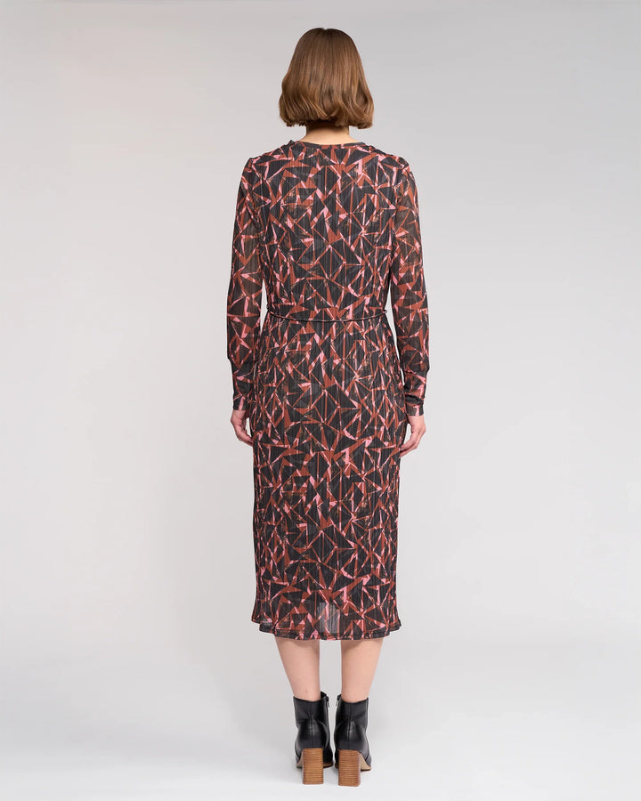 A woman with short light brown hair wears the Knewe Label Freddie Dress - Dusk, a long-sleeved, mid-length winter dress in red and black, paired with black block-heel ankle boots against a plain light background.