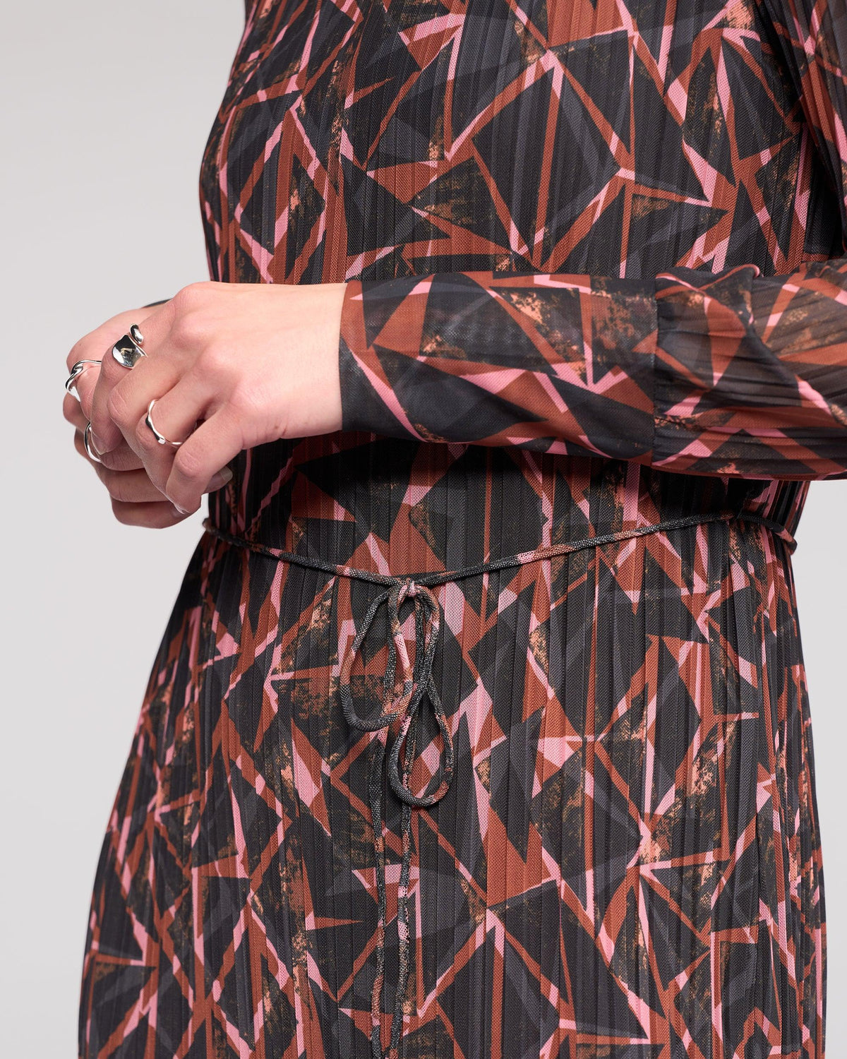 A close-up of someone wearing the Knewe Label Freddie Dress - Dusk, a crinkle mesh winter dress with geometric patterns in brown, black, and pink, featuring a waist tie. The person’s face isn’t visible and they are wearing rings.
