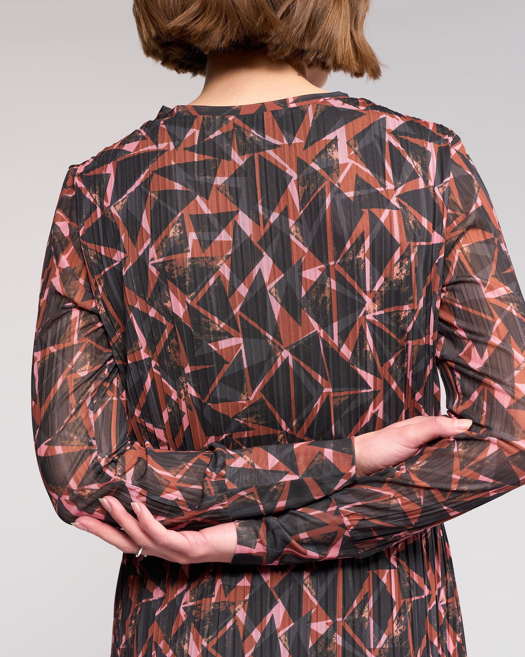 A person with short brown hair faces away from the camera, arms crossed behind them, wearing the Freddie Dress - Dusk by Knewe Label, a long-sleeved winter dress with a geometric pink and brown print.