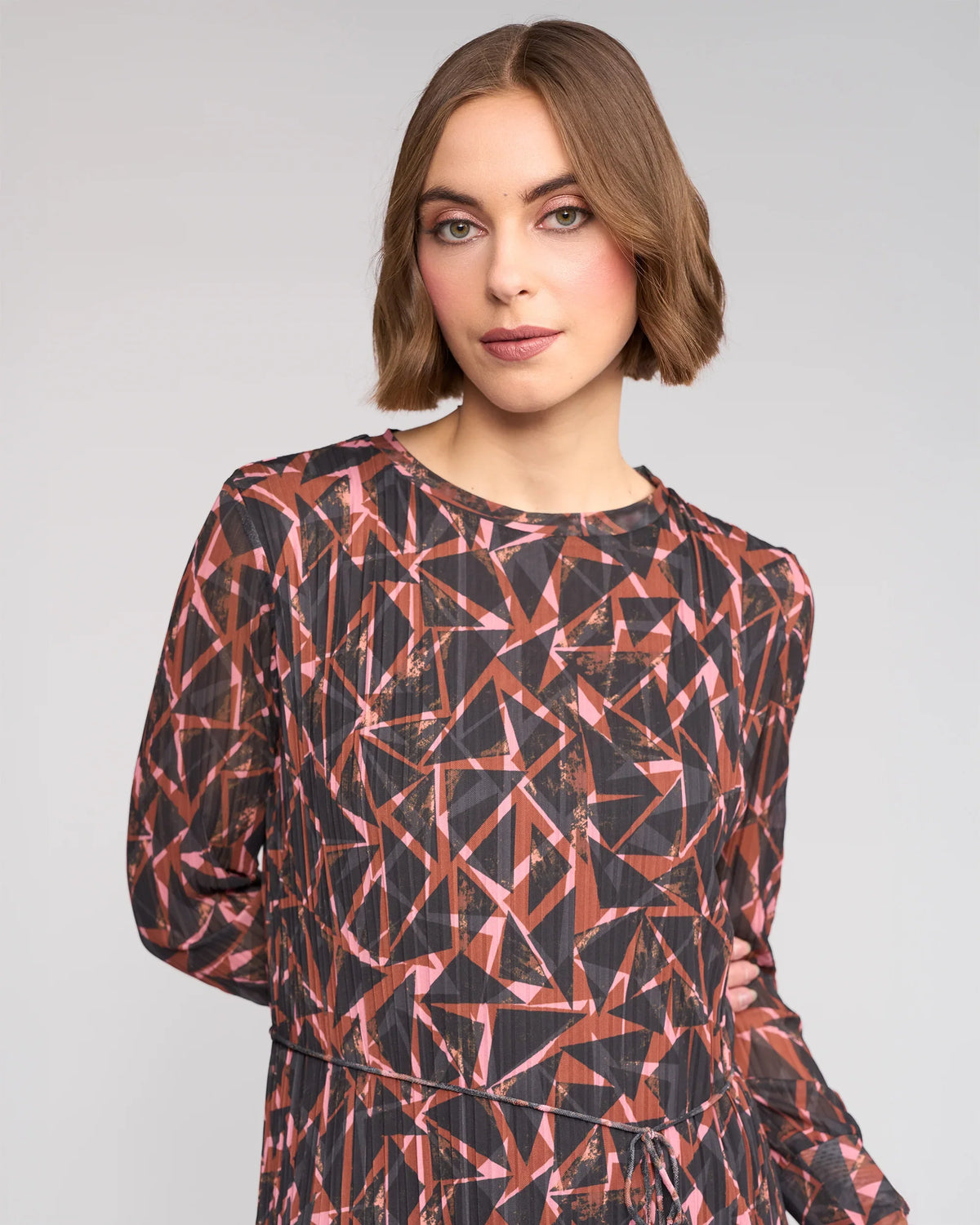 A woman with chin-length brown hair models the Knewe Label Freddie Dress - Dusk, a long-sleeved winter dress with a geometric dusk print in red, pink, and black, against a light gray background.