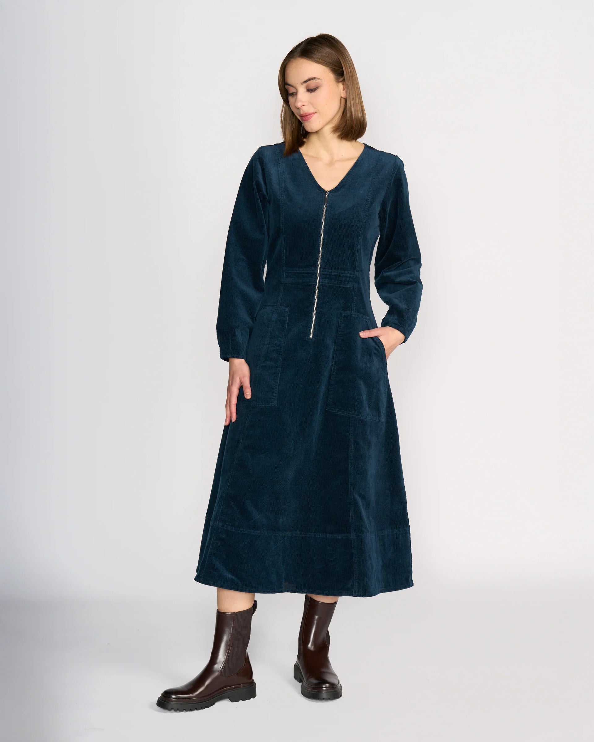 A woman stands against a plain background, wearing the Knewe Label Phoenix Dress - Airforce, a long sleeve dark blue cord dress with a front zipper, V-neckline, and pockets. She pairs it with dark brown ankle boots and has straight shoulder-length hair.