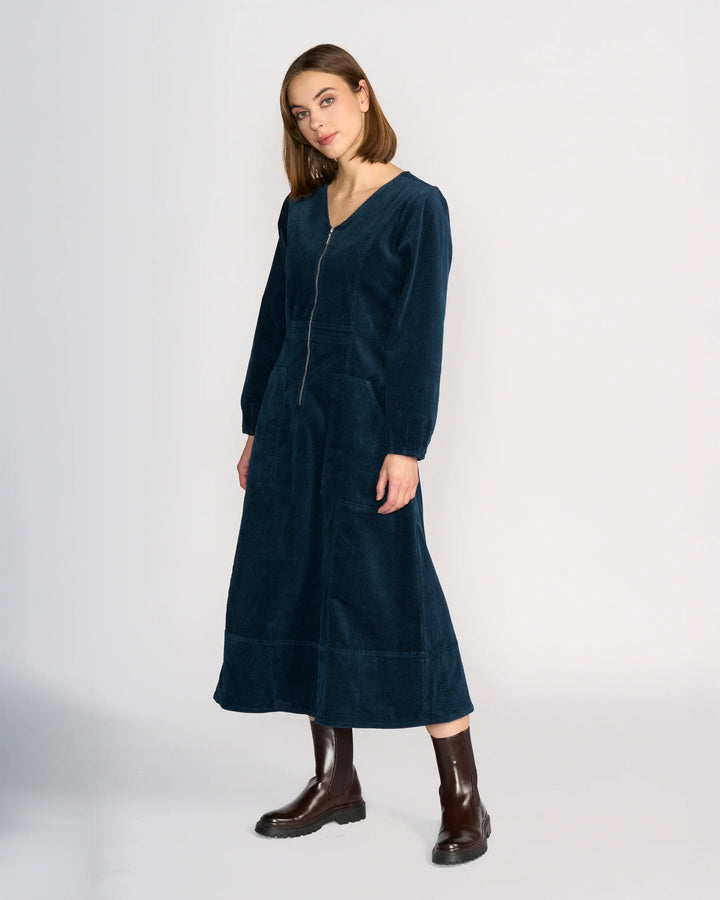 A woman with straight brown hair wears the Knewe Label Phoenix Dress in Airforce, a dark blue, long-sleeved cord dress with a zip front and V-neckline, paired with dark brown ankle boots against a plain light background.