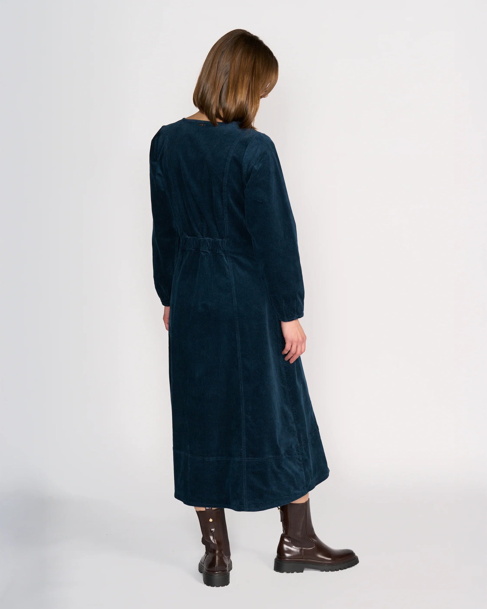 A person with straight, shoulder-length brown hair stands with their back to the camera, wearing the Knewe Label Phoenix Dress - Airforce and dark brown boots against a plain white background.