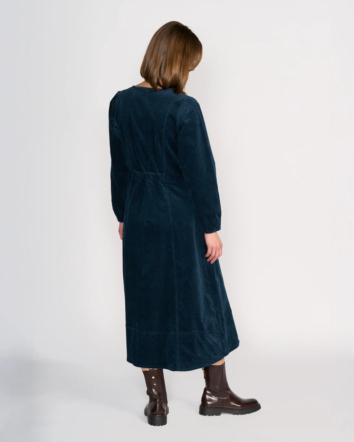 A person with straight, shoulder-length brown hair stands with their back to the camera, wearing the Knewe Label Phoenix Dress - Airforce and dark brown boots against a plain white background.