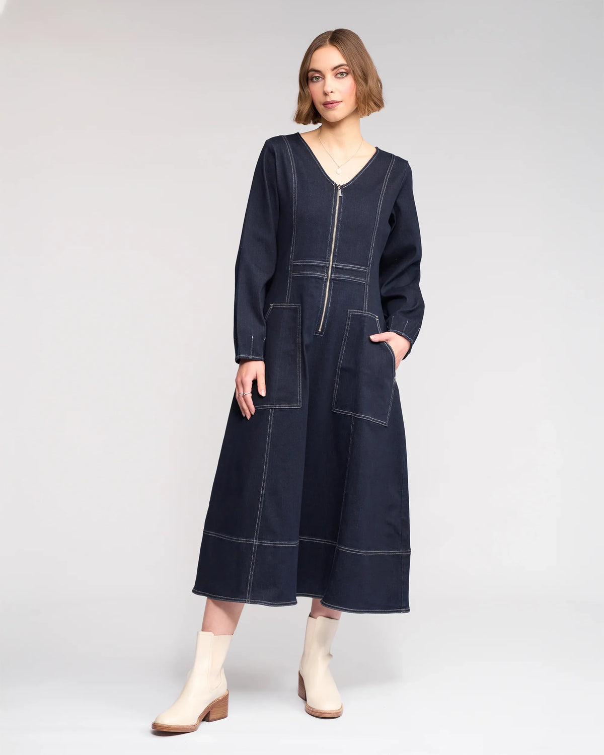 A woman models the Knewe Label Phoenix Dress - Ink Denim, featuring long sleeves, a V-neck, white stitching, front zipper, and large pockets. She completes the look with ankle-high white boots and one hand in her pocket.
