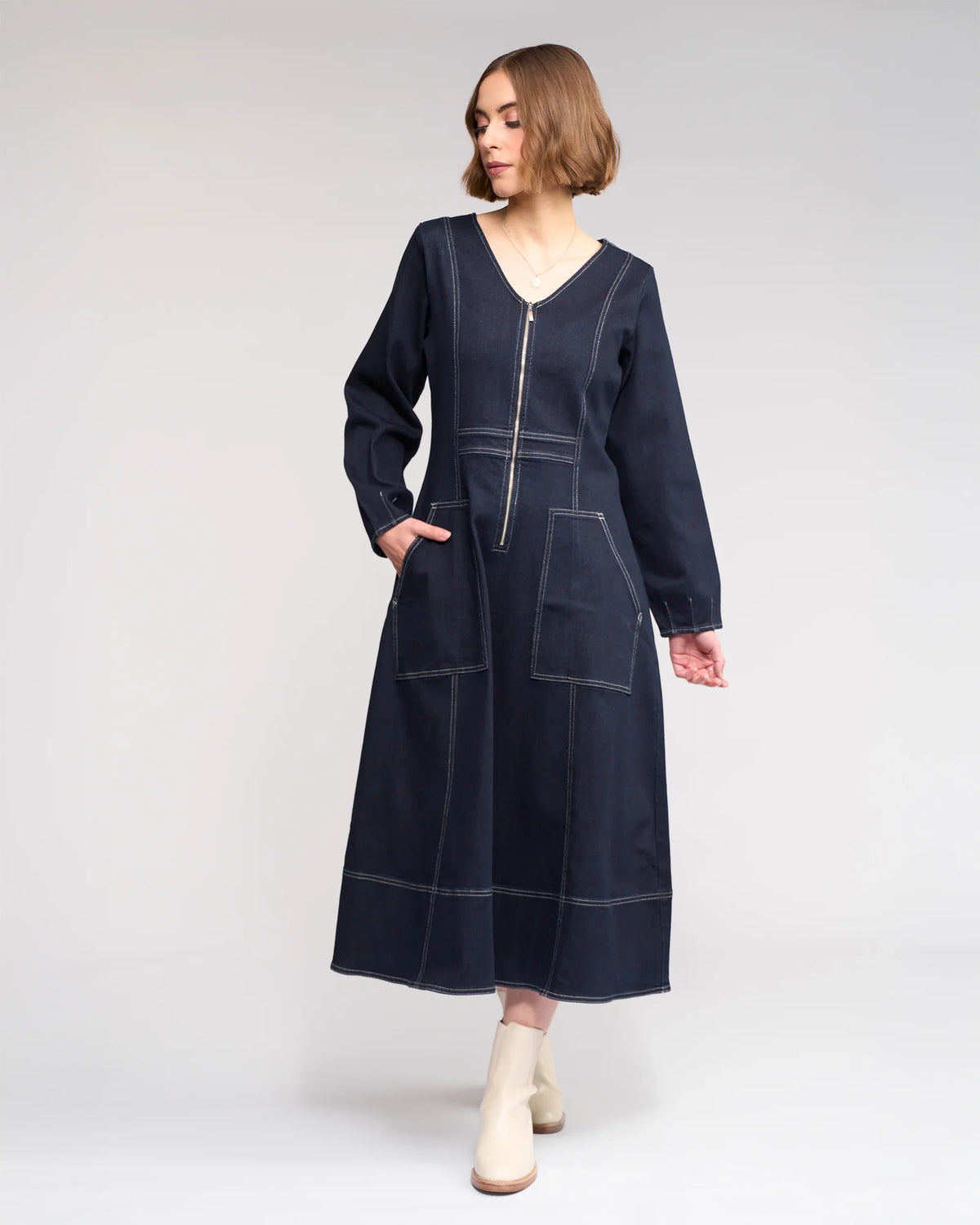A woman in the Phoenix Dress - Ink Denim by Knewe Label, featuring long sleeves, a V-neckline, contrast white stitching, patch pockets, and a front zipper, pairs it with cream ankle boots and stands against a light gray background.