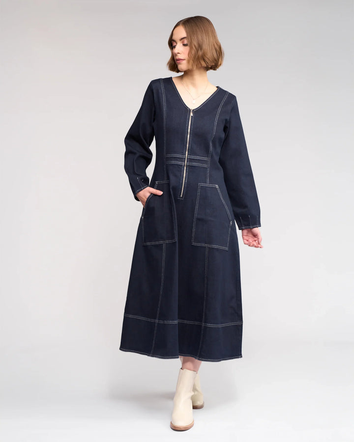 A woman in the Phoenix Dress - Ink Denim by Knewe Label, featuring long sleeves, a V-neckline, contrast white stitching, patch pockets, and a front zipper, pairs it with cream ankle boots and stands against a light gray background.
