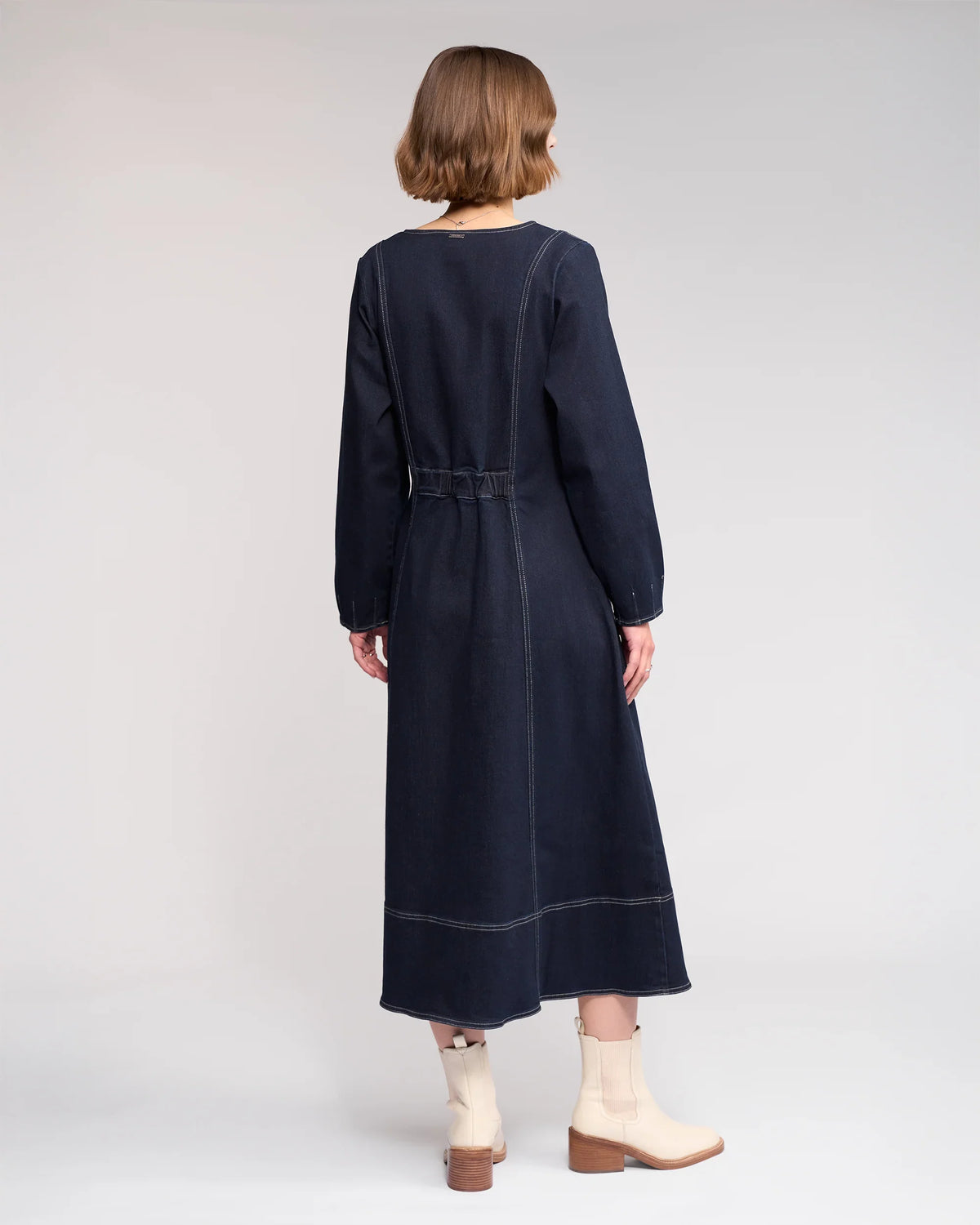 A woman with short, light brown hair faces away, wearing the Knewe Label Phoenix Dress in Ink Denim and light-colored ankle boots with wooden heels. The background is plain and light-colored.