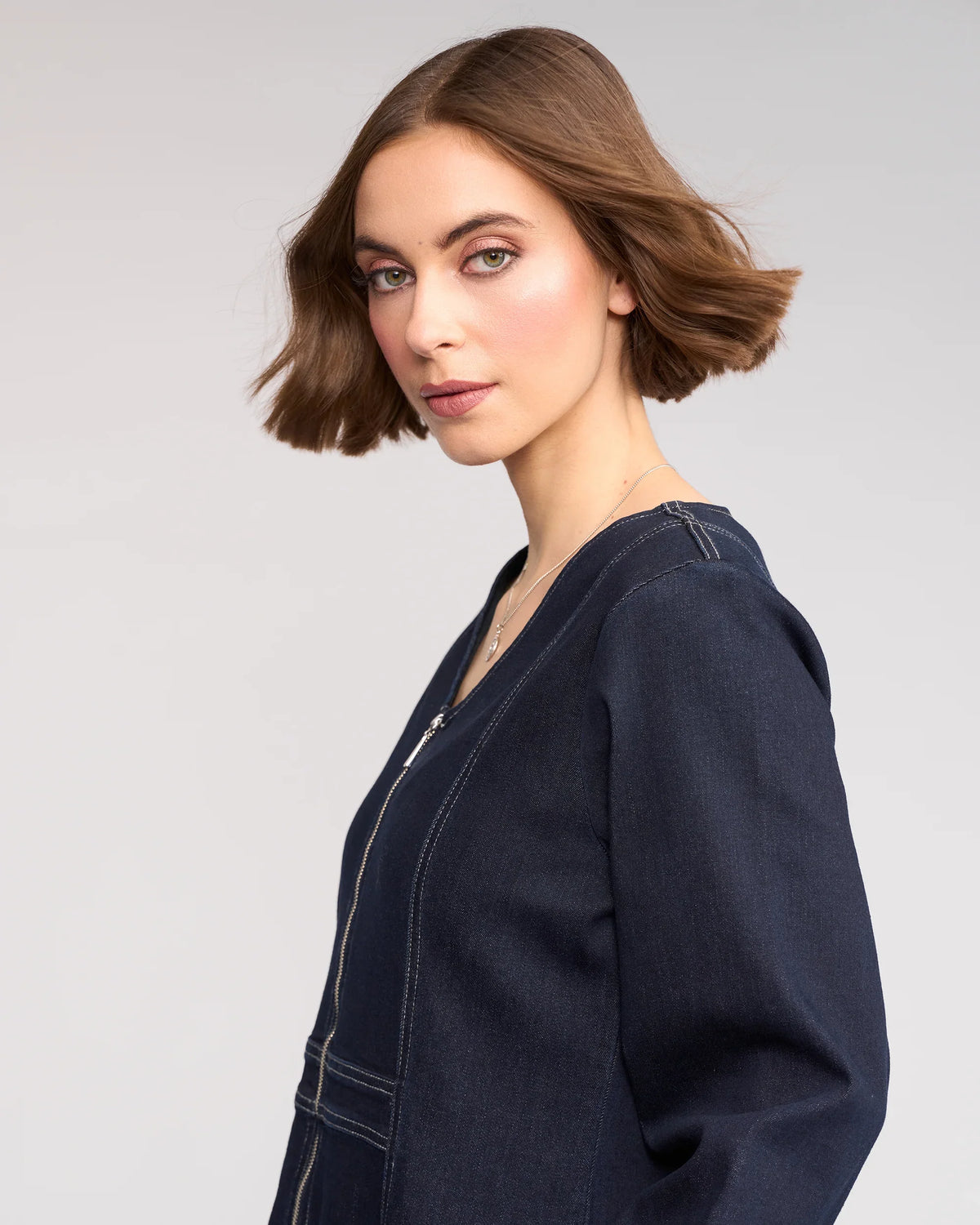 A woman with chin-length brown hair and light makeup poses against a plain background, wearing the Knewe Label Phoenix Dress in Ink Denim—a dark, long sleeve denim dress with white stitching and a subtle V-neckline. She looks over her shoulder at the camera.