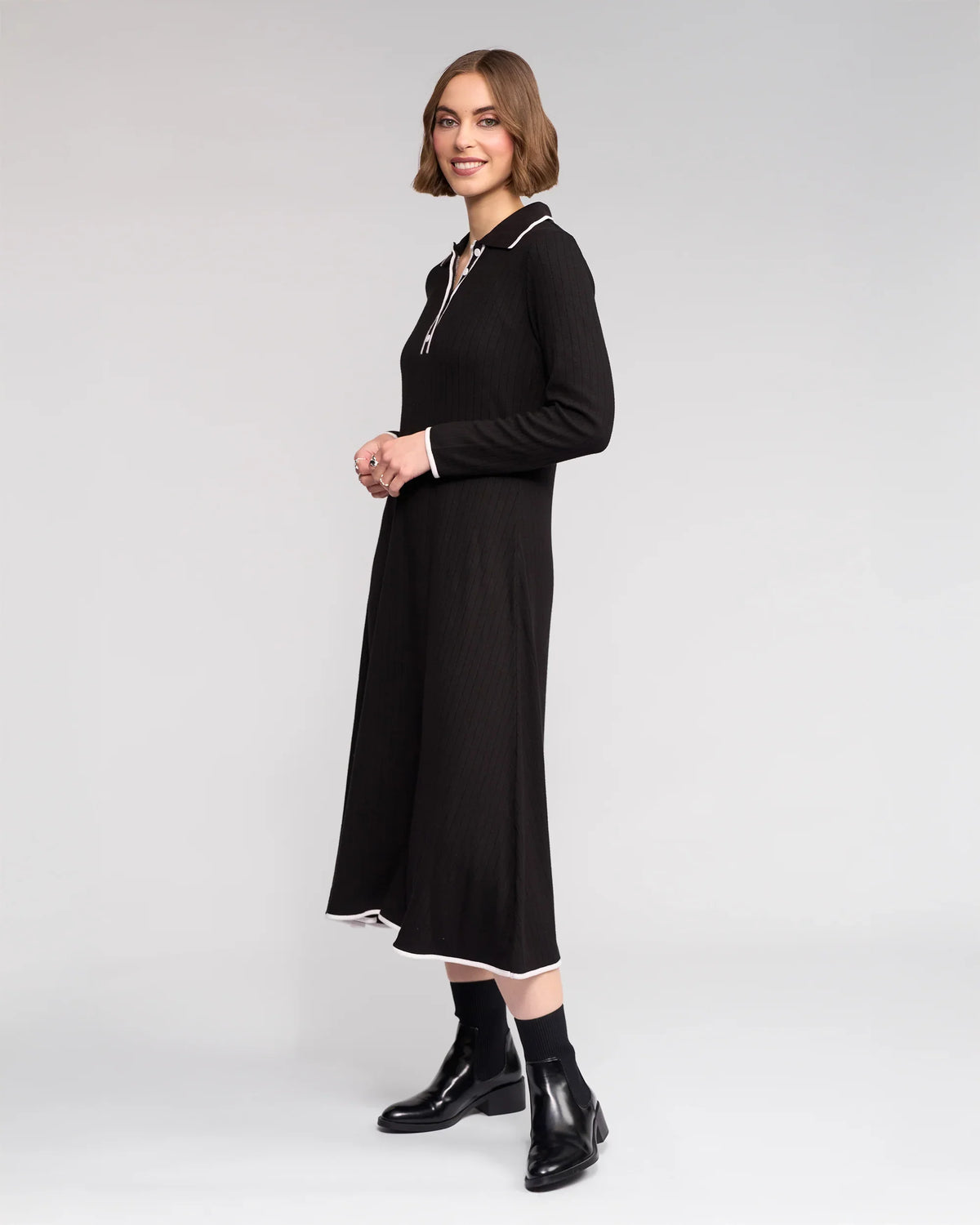 A woman with short brown hair wears the Knewe Label Ritual Dress in black, featuring long sleeves and white trim on the collar and cuffs. She pairs it with black ankle boots, standing sideways and smiling against a plain light background.