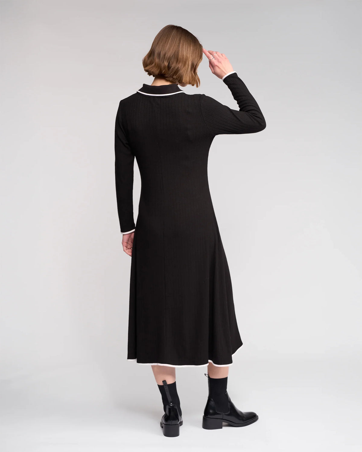 A person with short hair, seen from behind against a plain light backdrop, wears the Knewe Label Ritual Dress in black featuring a white shirt-style collar and cuffs, paired with black ankle boots.