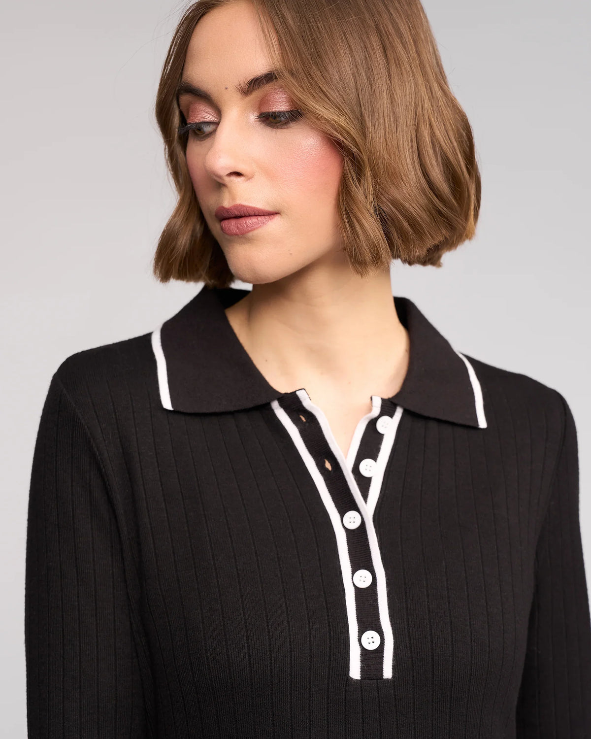 A woman with short, light brown hair looks down to her left, wearing the Knewe Label Ritual Dress in black—a ribbed knit style with a shirt collar, white trim, and white buttons. The background is light gray.